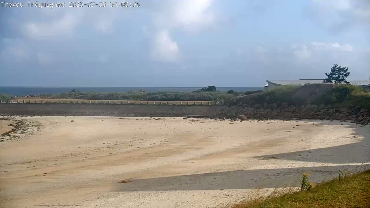 A sandy beach stretches out to a calm ocean under a partly cloudy sky, with a building and some vegetation visible in the background.