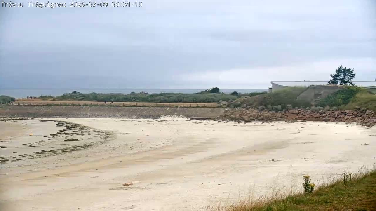 A sandy beach stretches out to a calm sea under an overcast sky, with a low-lying structure visible on the far side.
