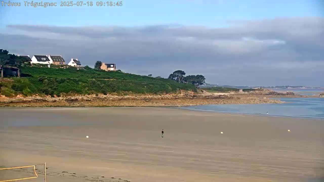 A sandy beach stretches out to calm water, backed by a grassy hillside with houses and a few trees under a partly cloudy sky.