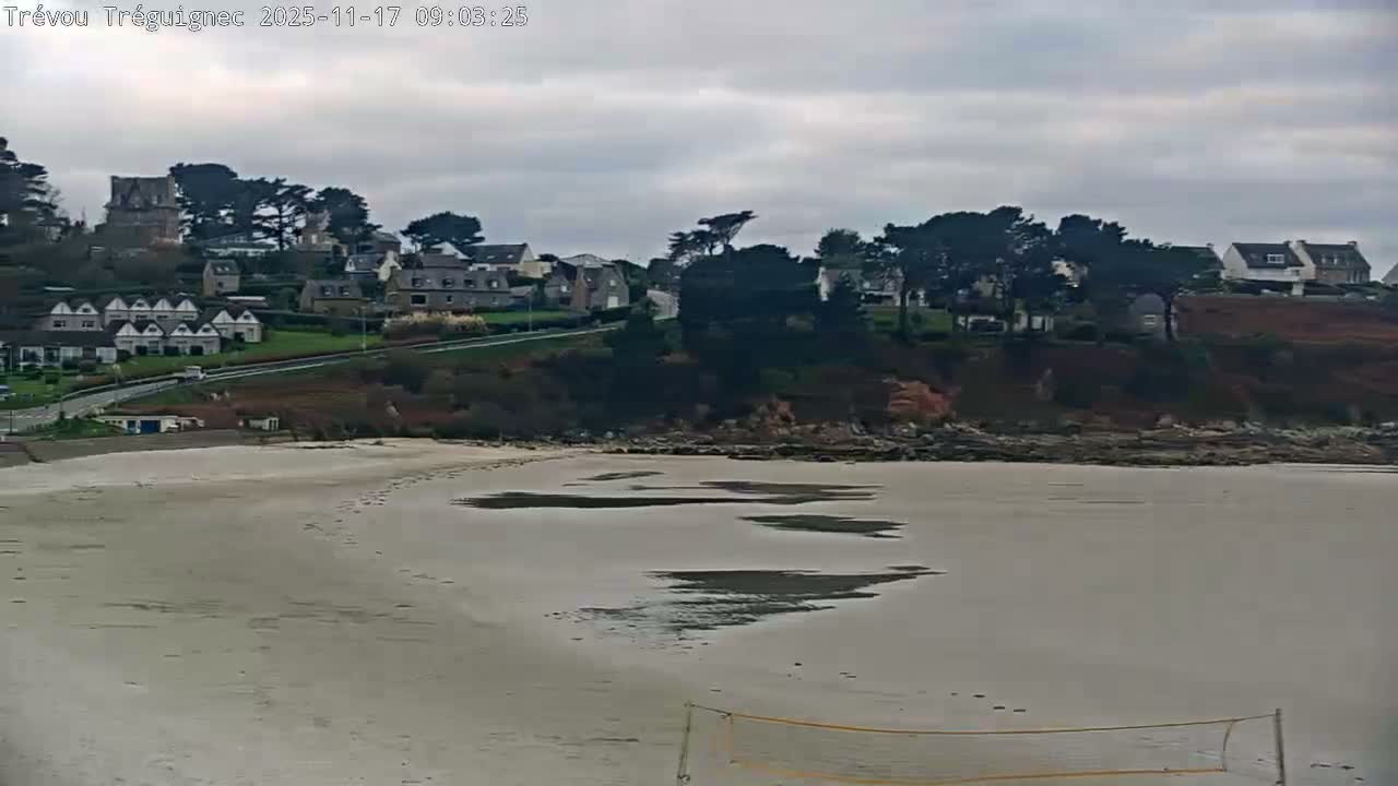 An overcast sky hangs over a coastal town featuring houses on a verdant hill, a road winding along the shore, and a sandy beach with tidal pools and a volleyball net in the foreground.