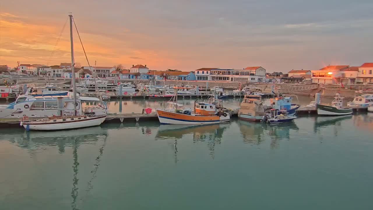 Many boats are moored in a harbor at sunset, with calm water reflecting the orange and pink sky.