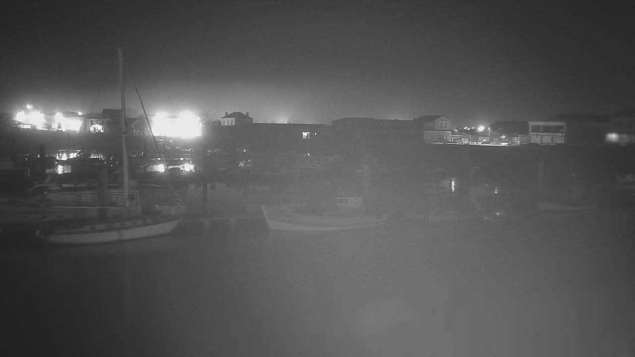 A sailboat is moored in a harbor at night, with buildings and lights faintly visible in the background.