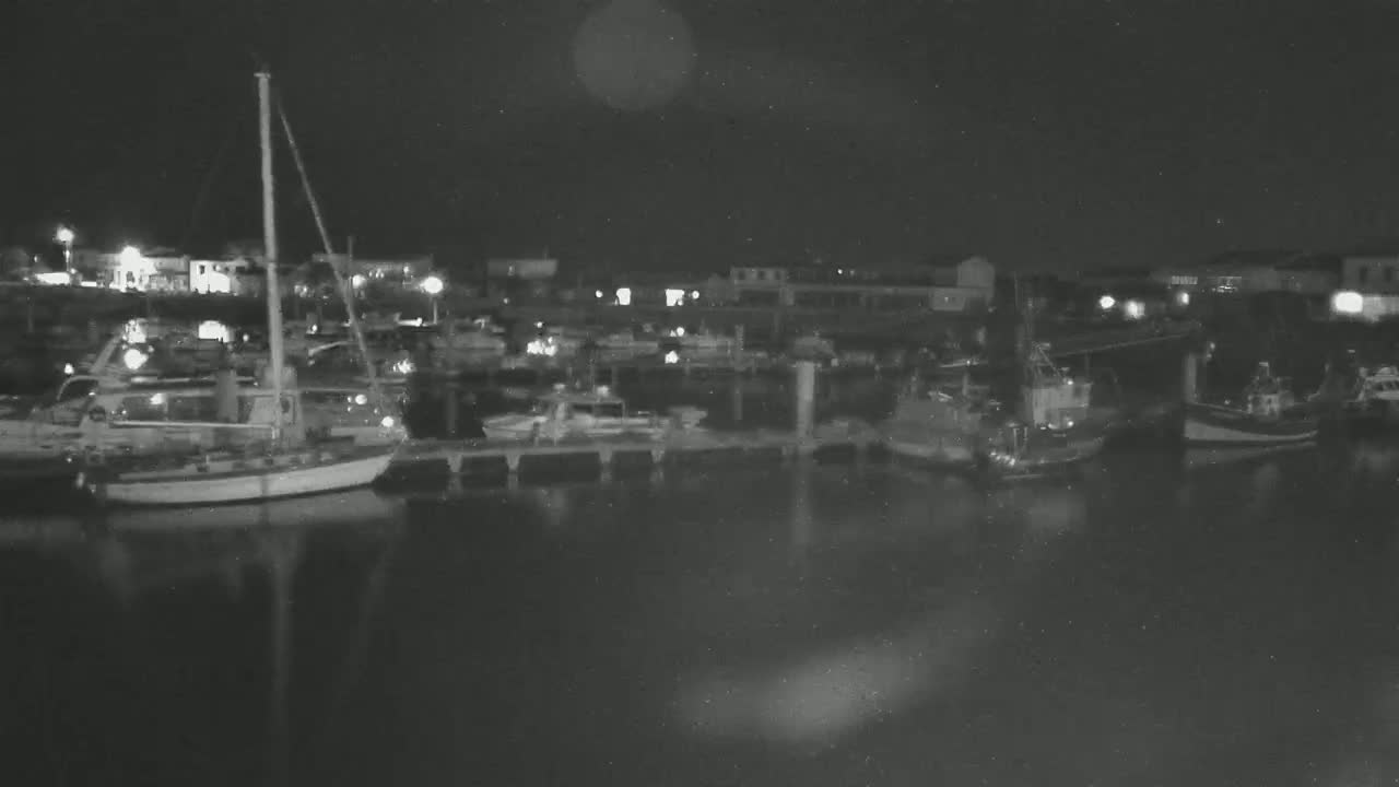 At night, a calm harbor is filled with numerous boats and sailboats moored at docks, some lights illuminating the scene.