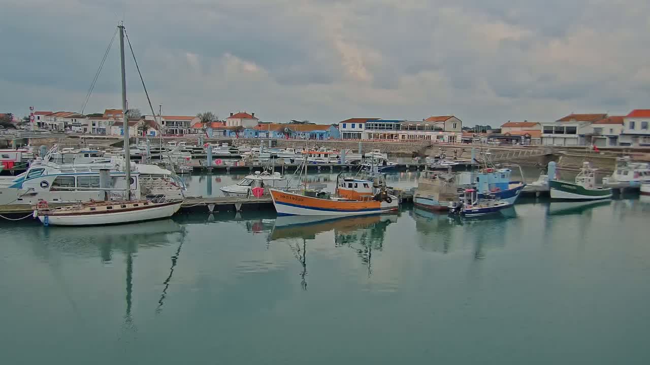 A calm harbor filled with sailboats and fishing vessels is nestled beside a waterfront town under a cloudy sky.