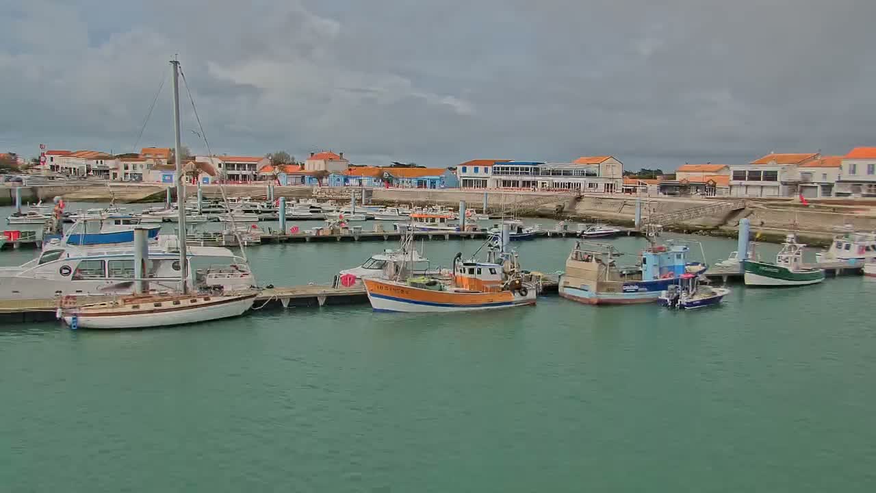 A harbor filled with various boats and yachts is nestled beside a waterfront town under a cloudy sky.