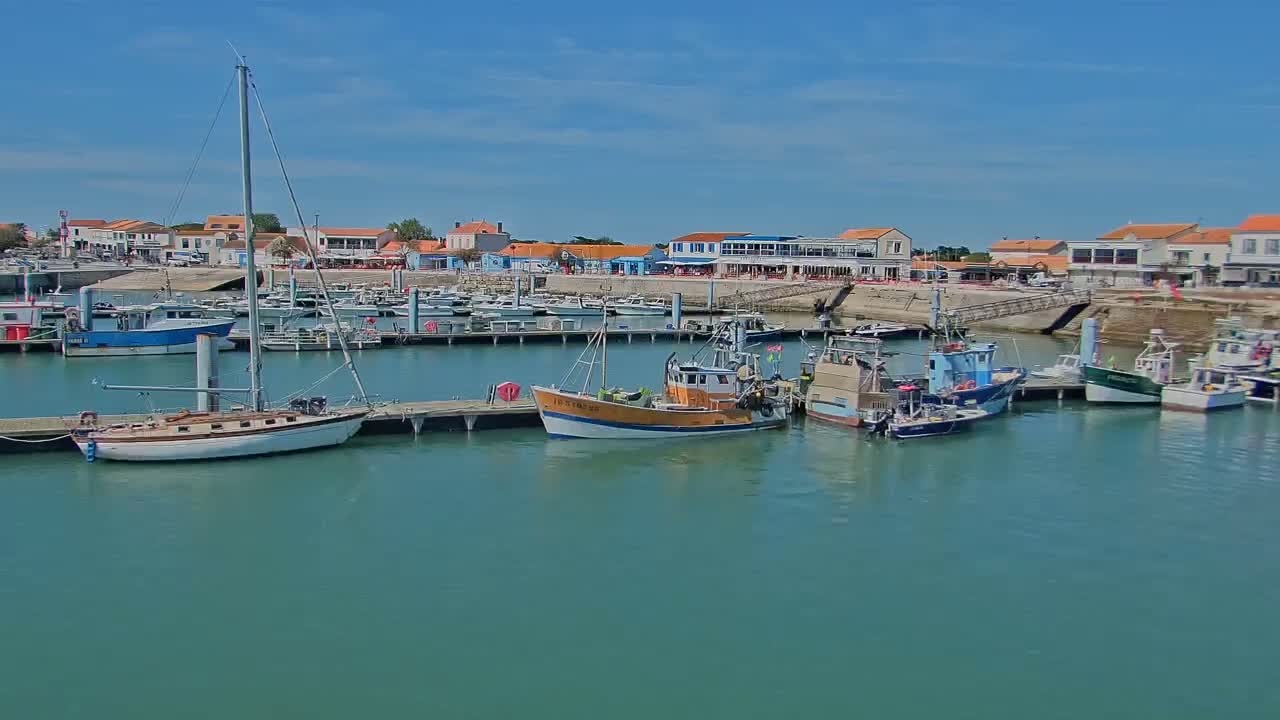 A marina filled with sailboats and fishing vessels is situated in front of a waterfront town under a clear blue sky.