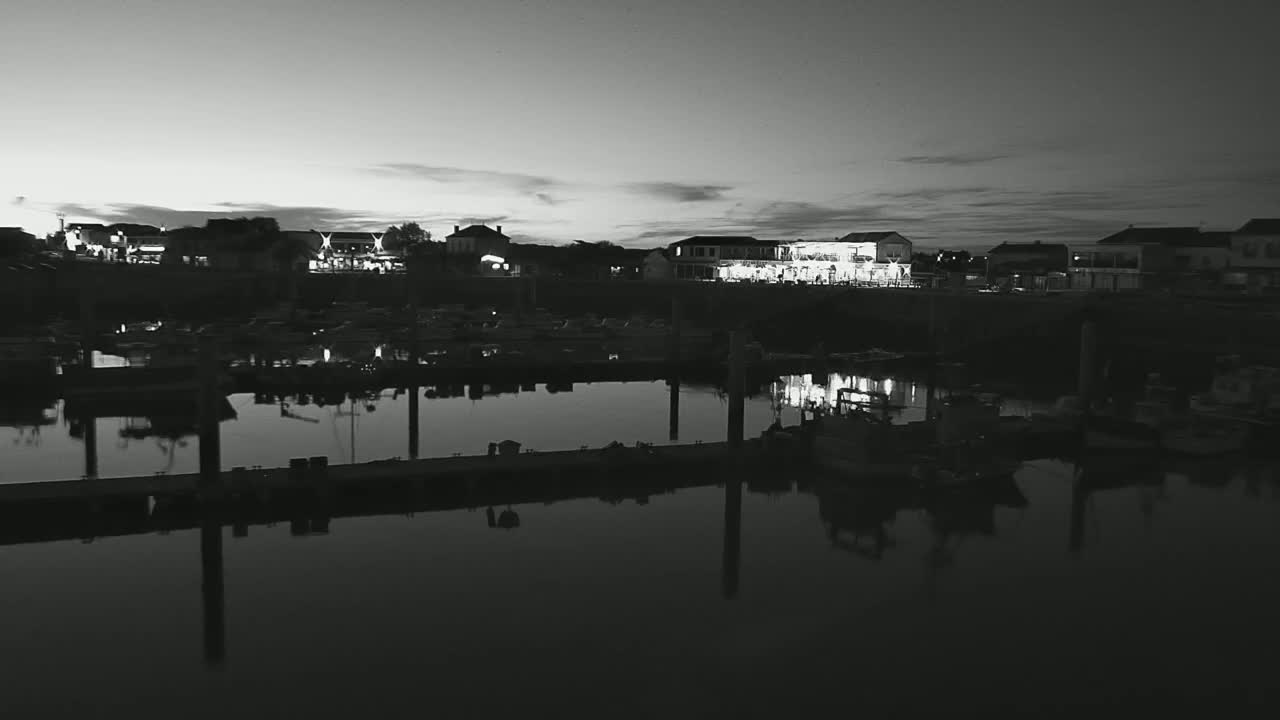 A nighttime, black-and-white view of a marina with buildings and boats reflecting in calm water under a dusky sky.