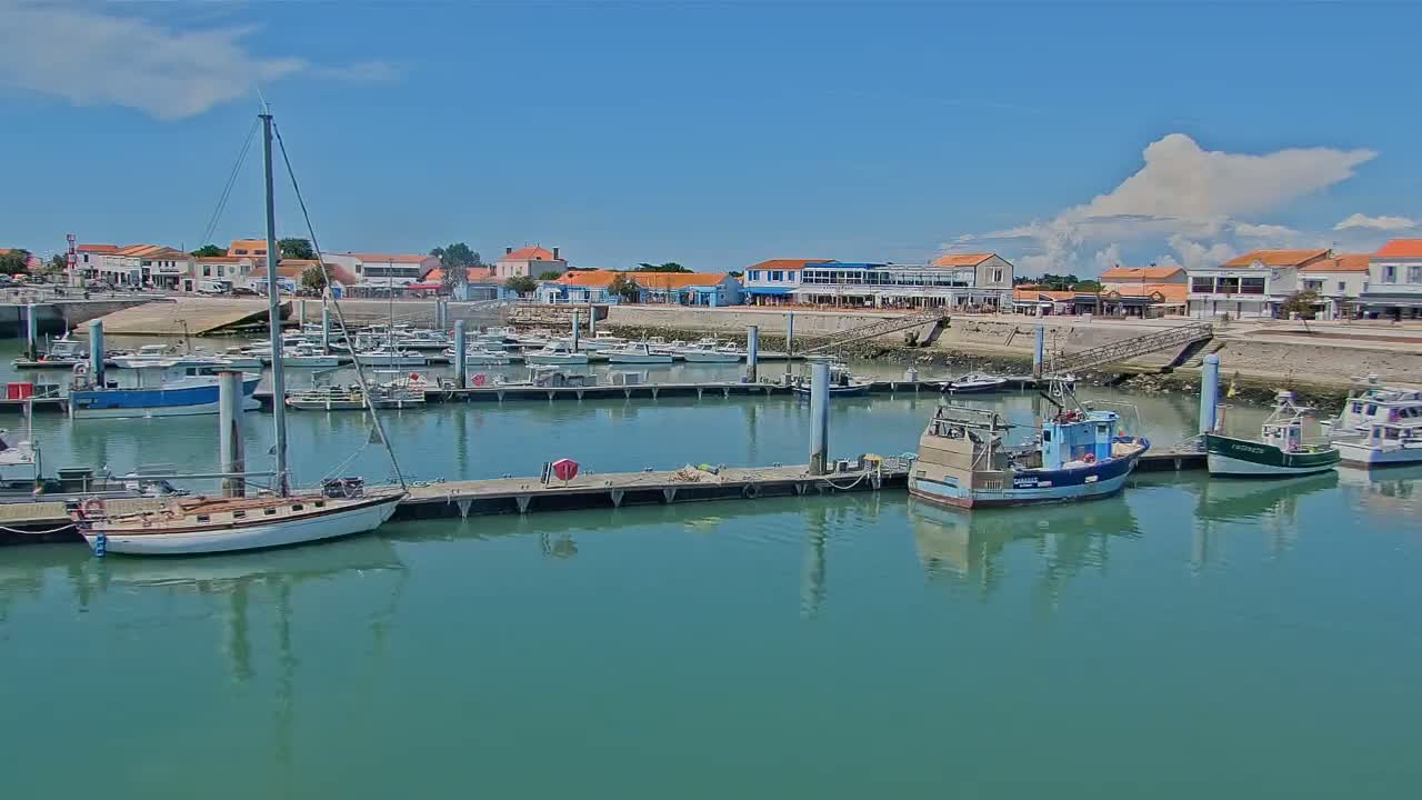 A marina filled with sailboats and fishing boats is situated in front of a waterfront town under a mostly sunny sky with some puffy clouds.