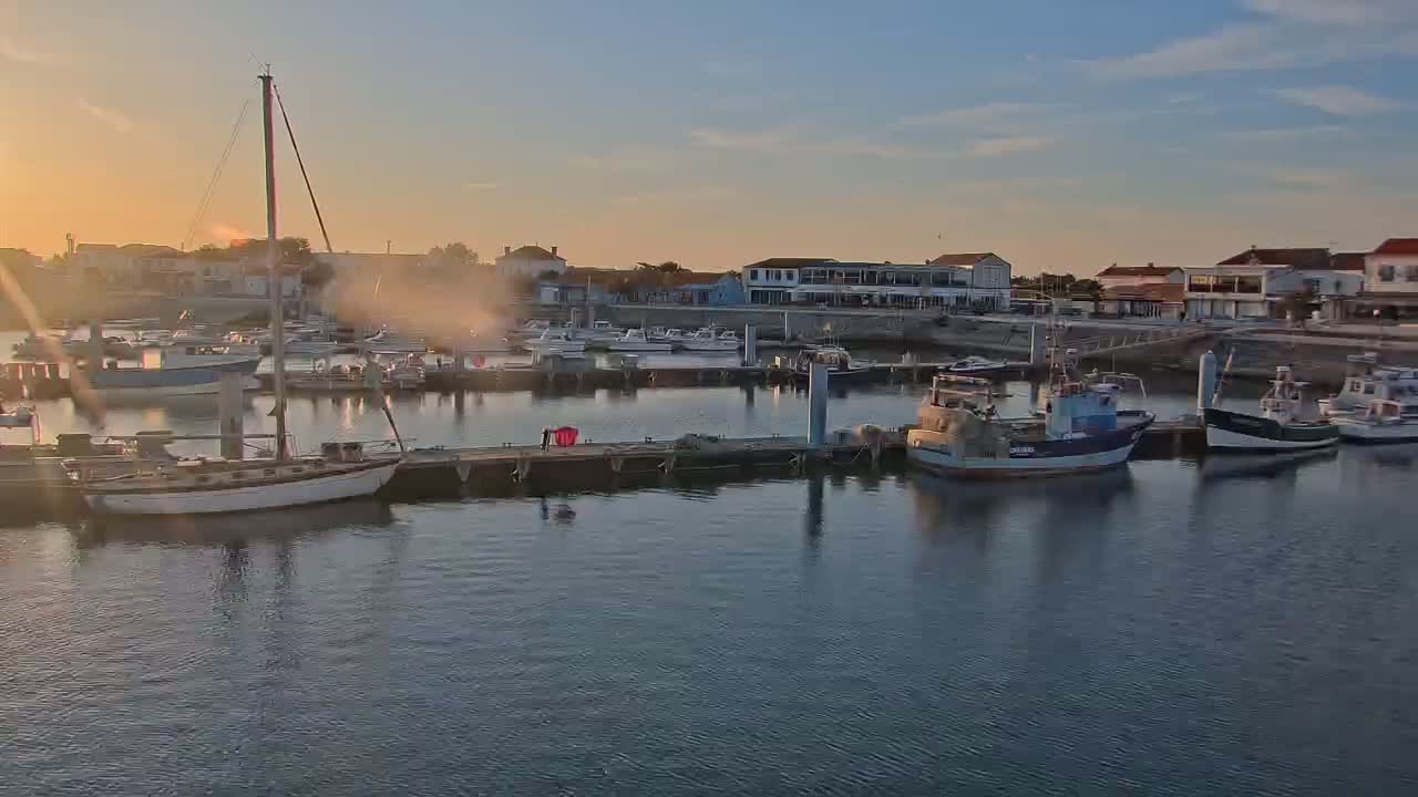A calm harbor at sunset, filled with sailboats and fishing vessels docked along piers beside a waterfront village.