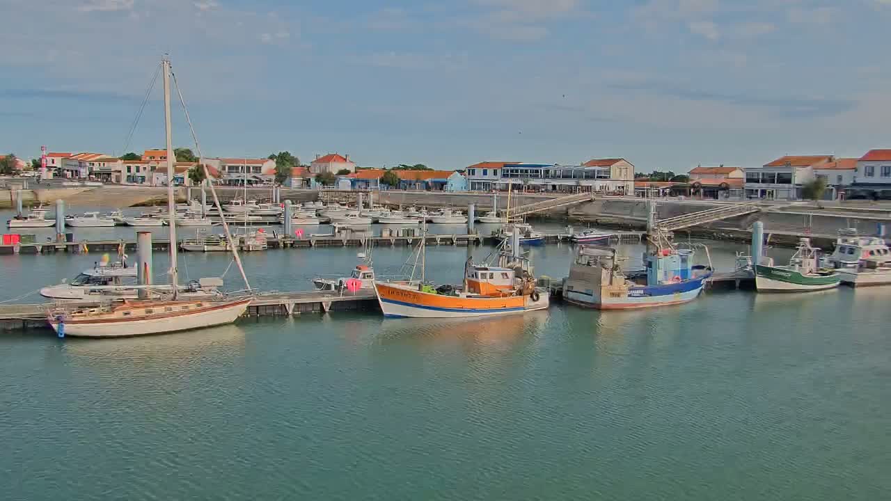 A marina filled with sailboats and fishing boats is situated in front of a waterfront town under a partly cloudy sky.