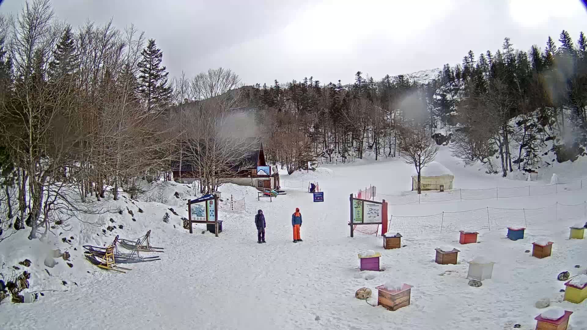 A snowy, overcast day at a winter recreation area shows a small building, two people standing near information boards, several snow sleds, and colorful storage boxes scattered across a snow-covered field surrounded by leafless trees.