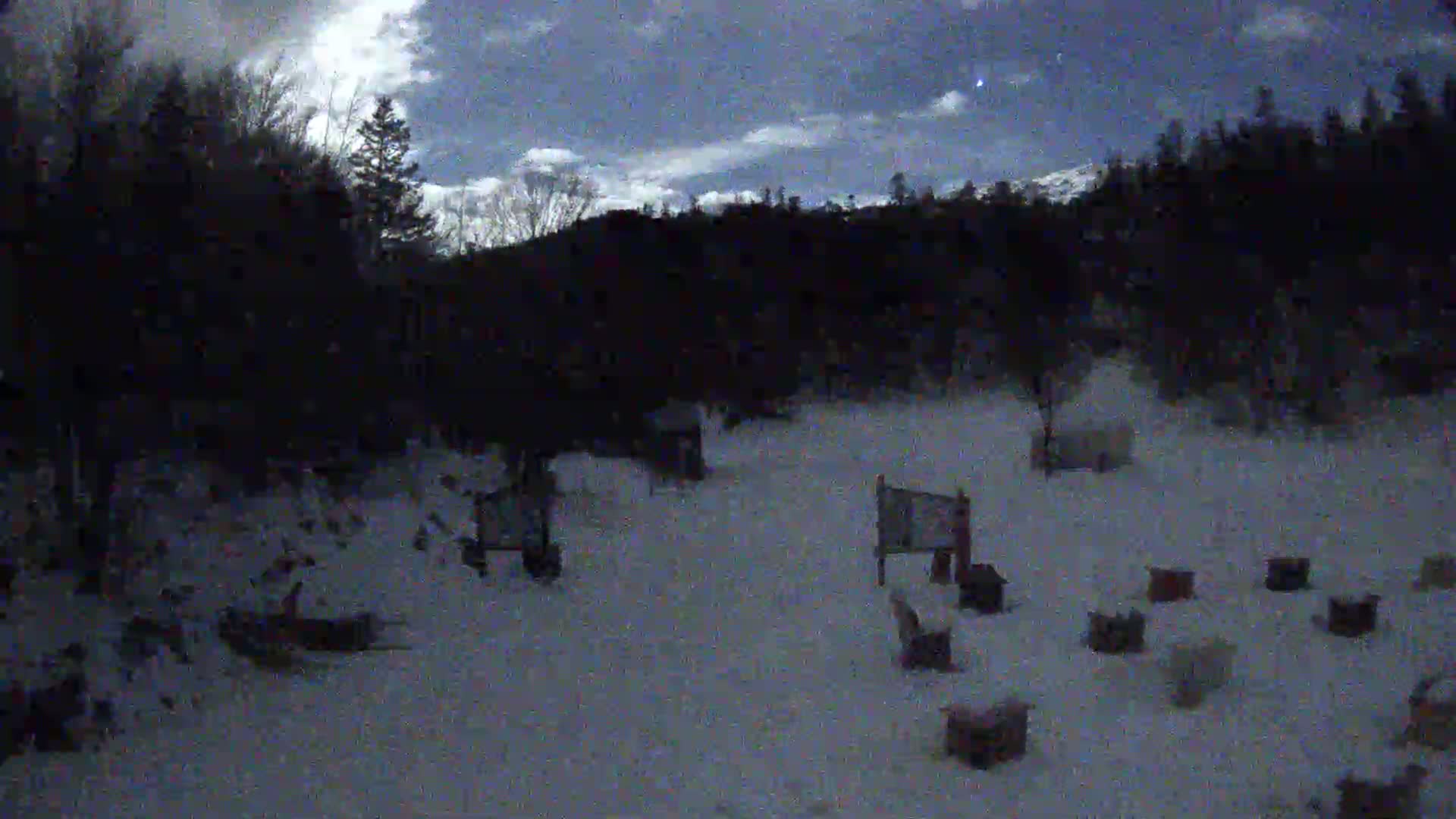 A snow-covered area at night, under a partly cloudy sky, shows several small structures scattered amidst dark trees and a hillside.