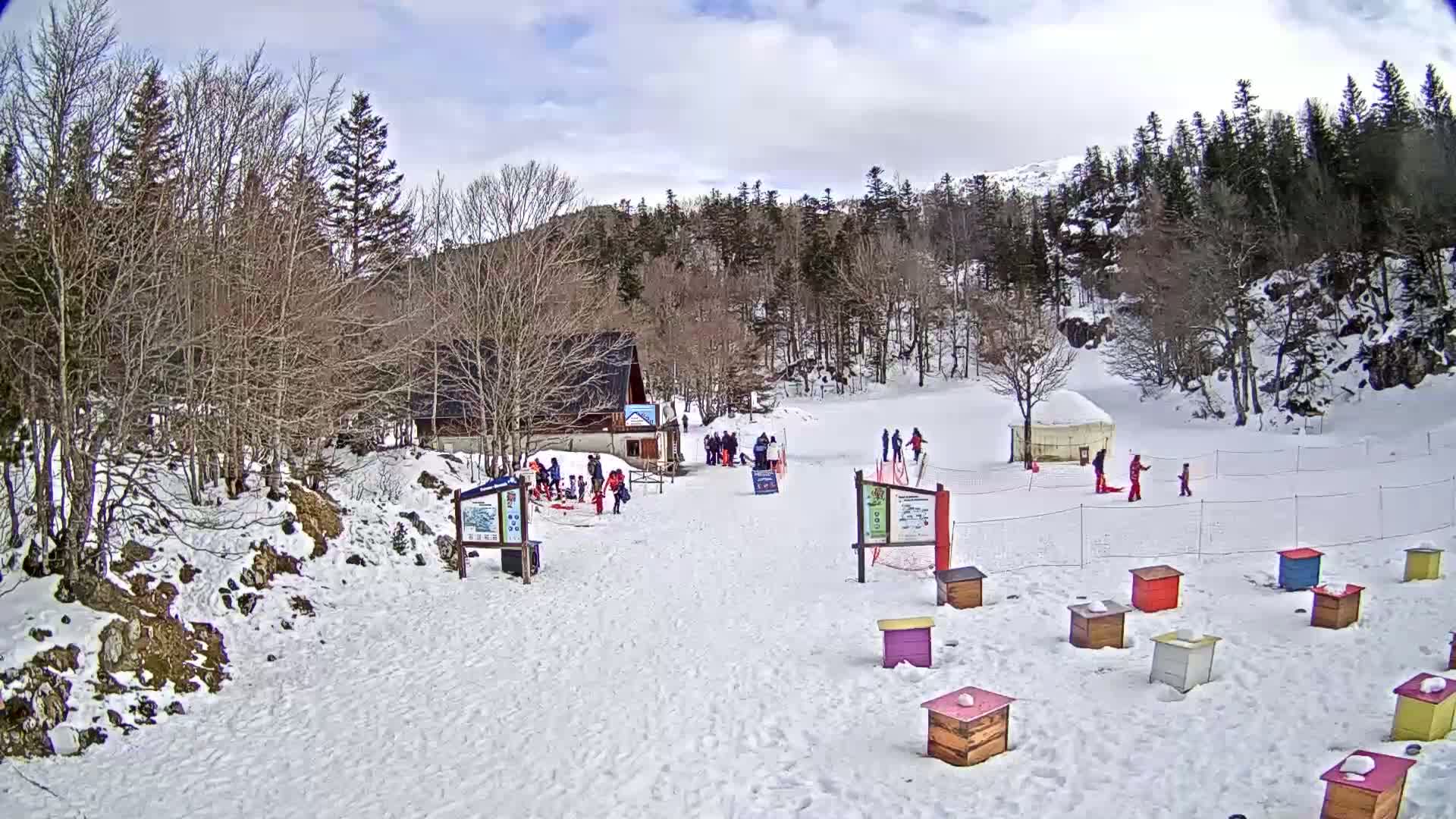 A snowy landscape with a small building, several people, a yurt, and colorful beehives under a partly cloudy sky.