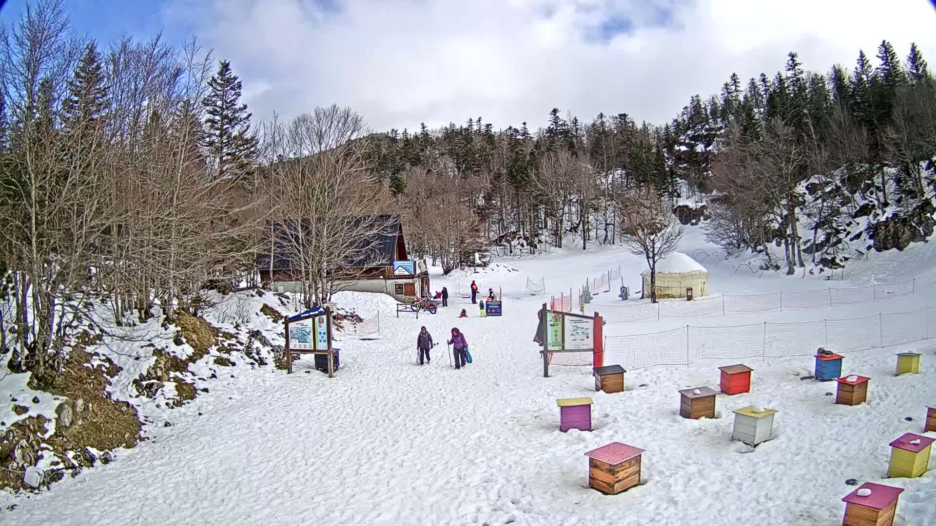 A snowy, lightly clouded day at a small ski slope with a few people and colorful beehives near a lodge surrounded by leafless trees.