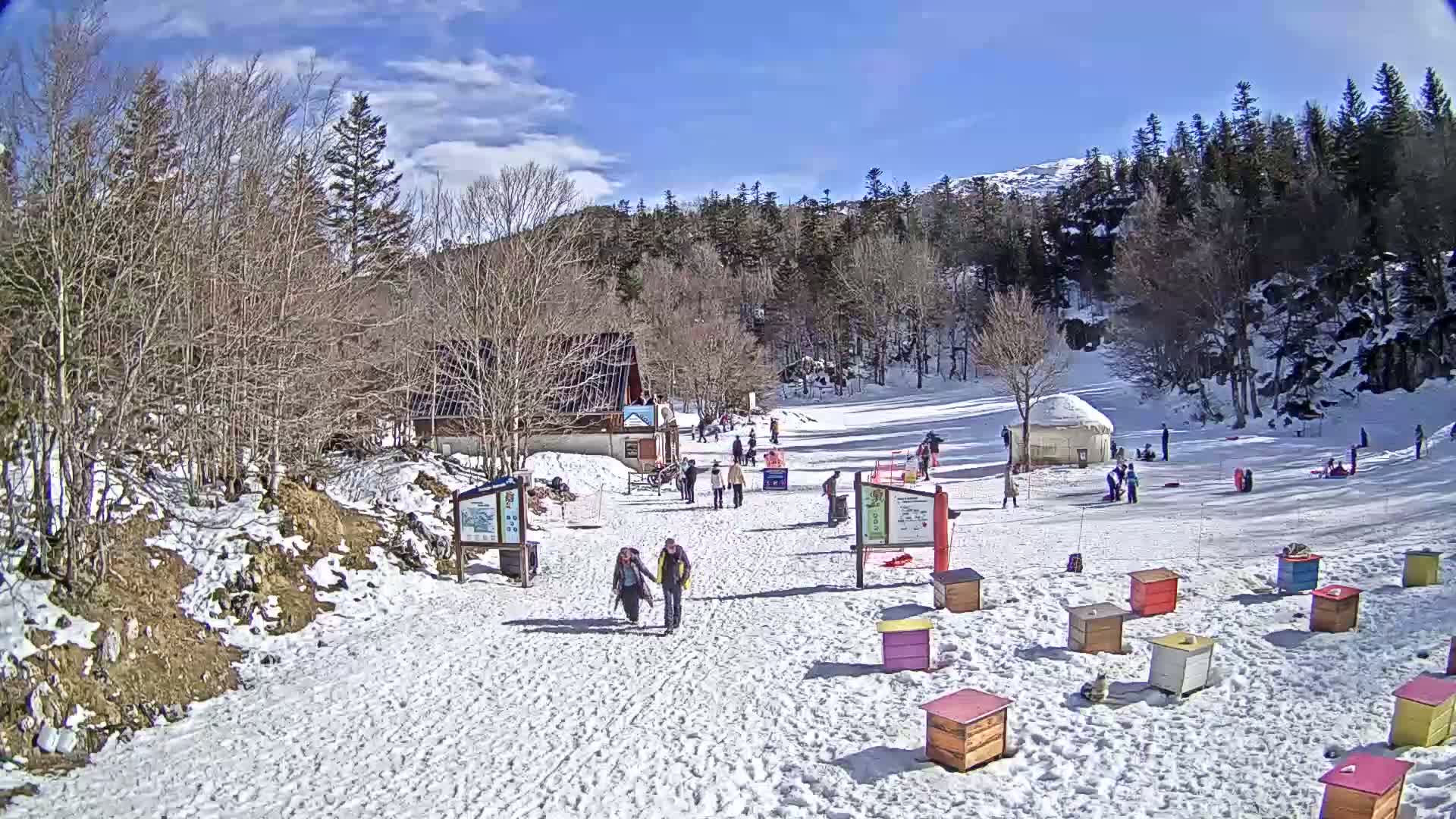 A snow-covered hill with a small building and several people, some sledding, under a sunny sky.