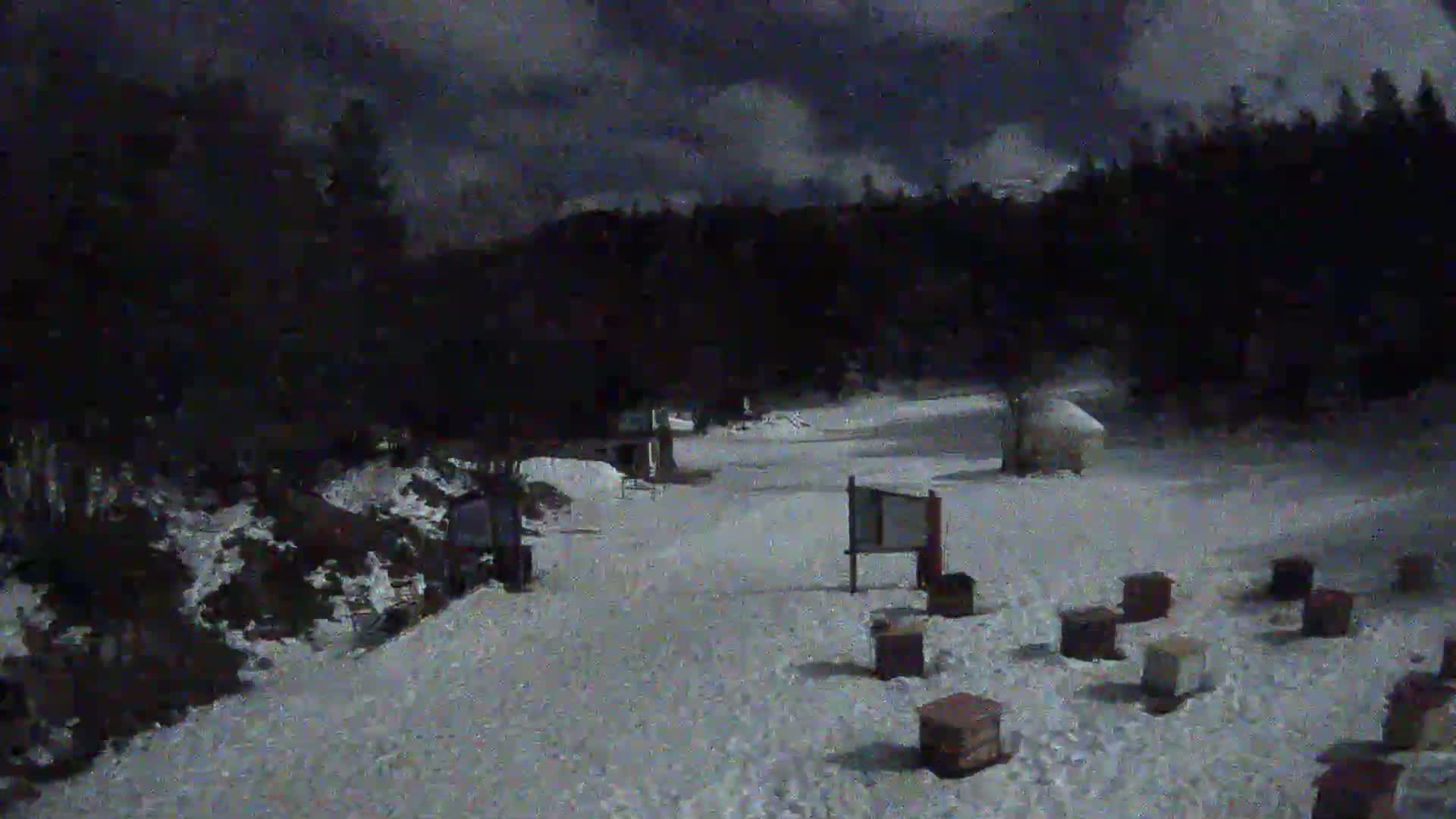 A snow-covered area with several small wooden structures and signage is illuminated under a dark, cloudy night sky.