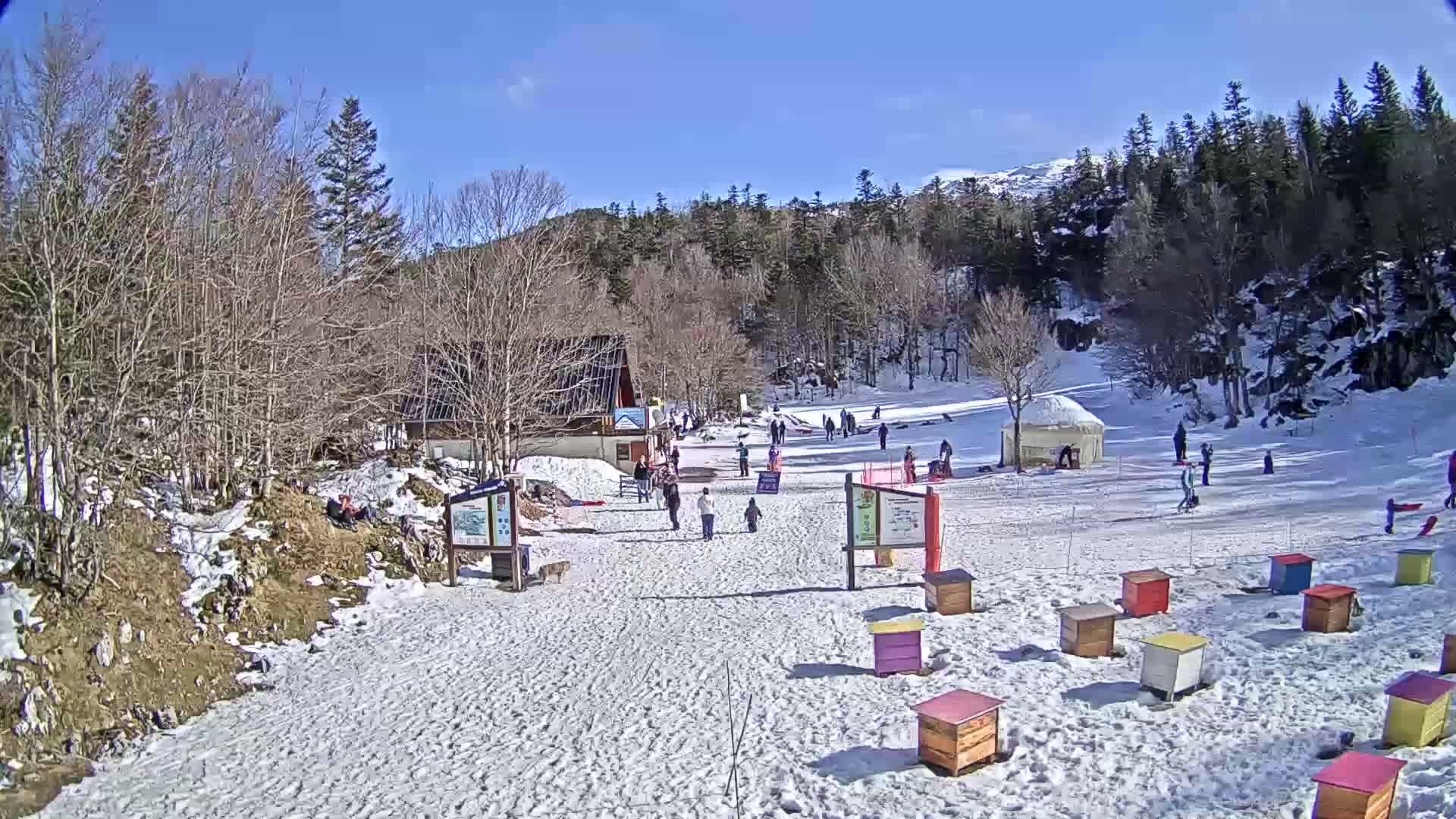 A snowy ski slope with a small building, several people, and colorful beehives under a sunny sky.