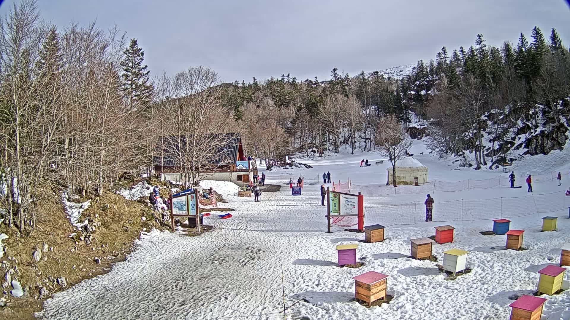 A snowy hilltop recreational area with a small building, a yurt, people enjoying winter activities, and a row of colorful beehives under a mostly sunny sky.