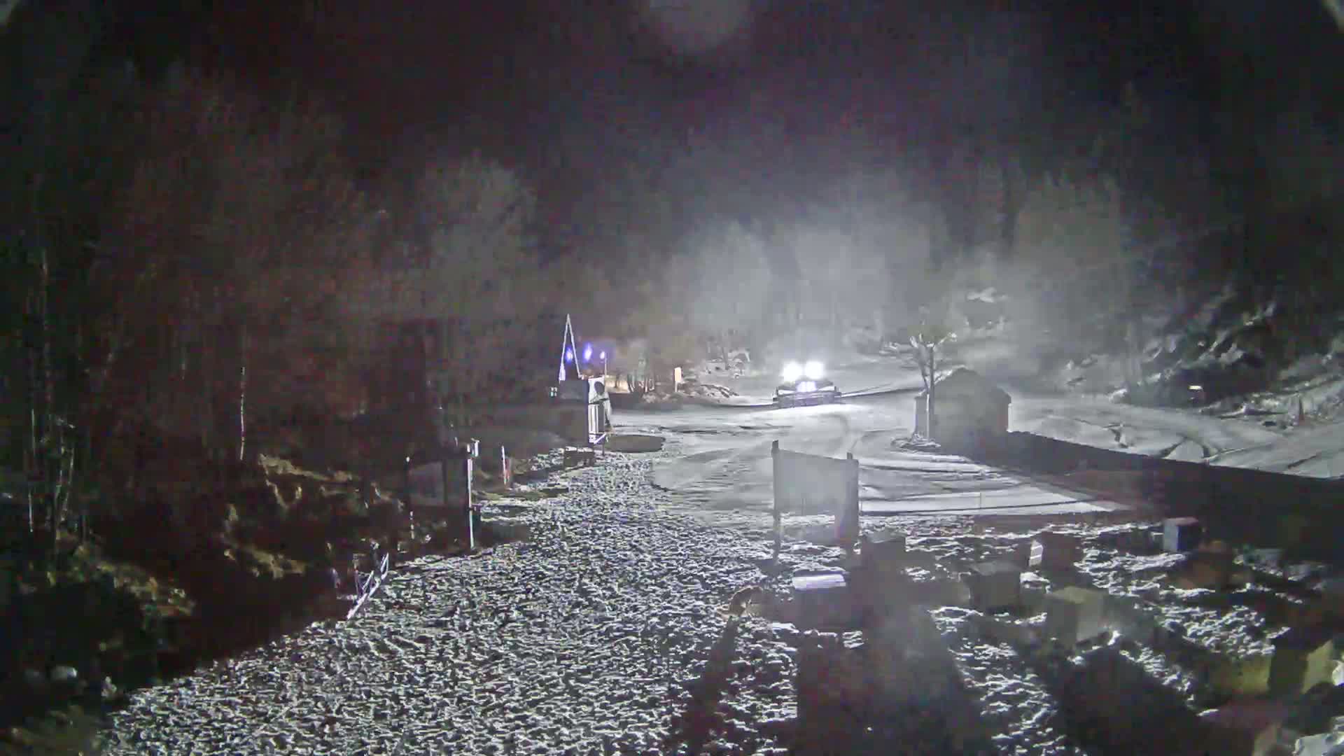 A snowcat drives down a snow-covered road at night, under hazy conditions.