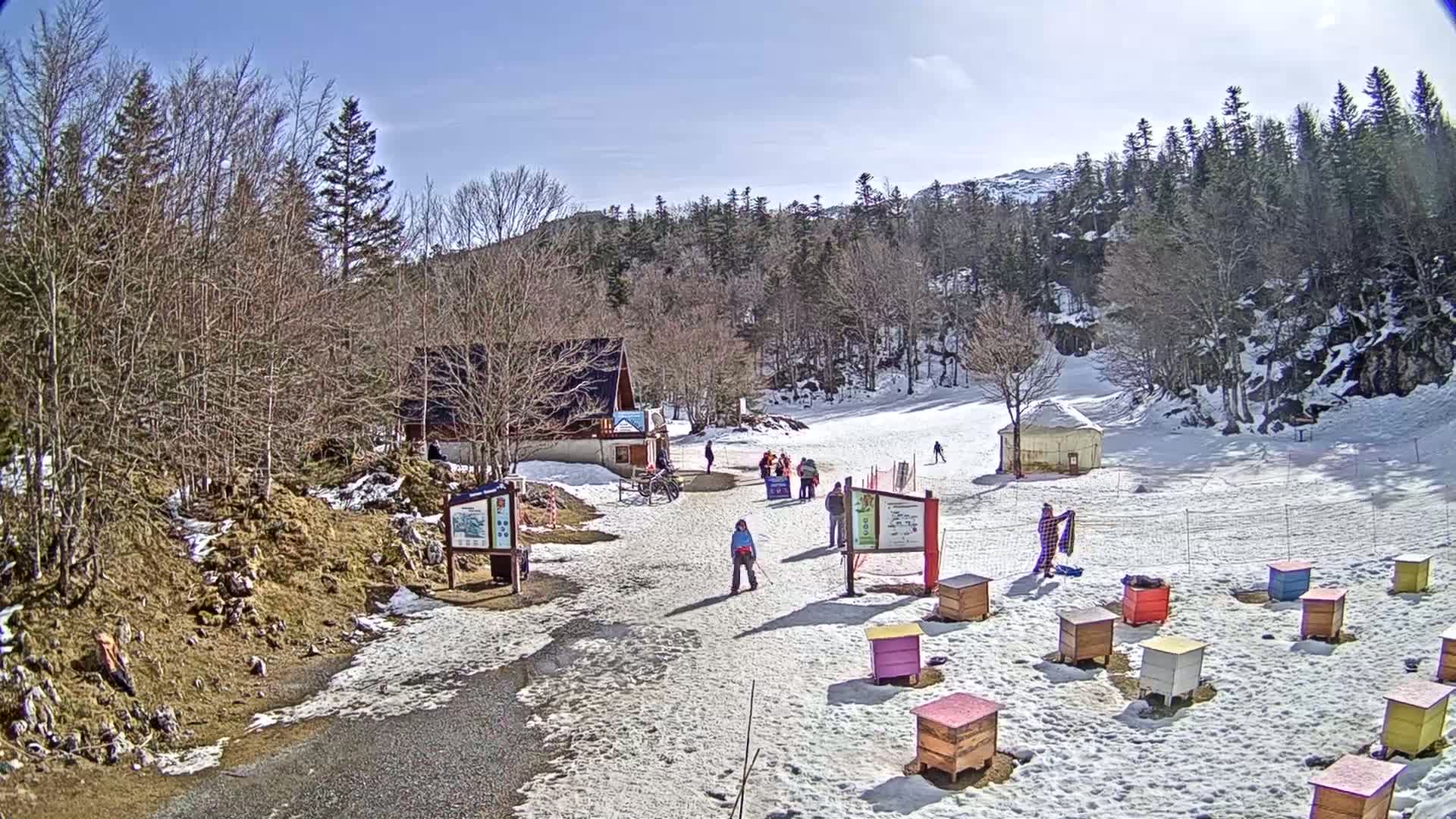 A sunny day at a snow-covered ski area shows a small building, several people, and numerous colorful beehives scattered across the snow.