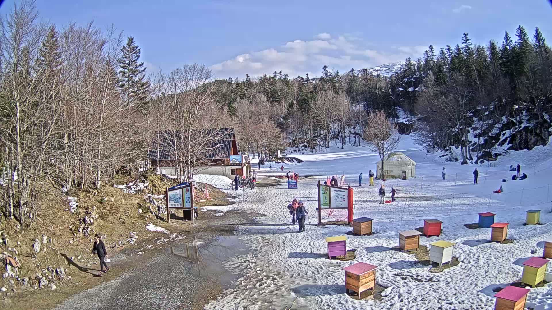A snowy, sunny scene shows a small ski area with a few people scattered about, a yurt, and numerous colorful beehives.