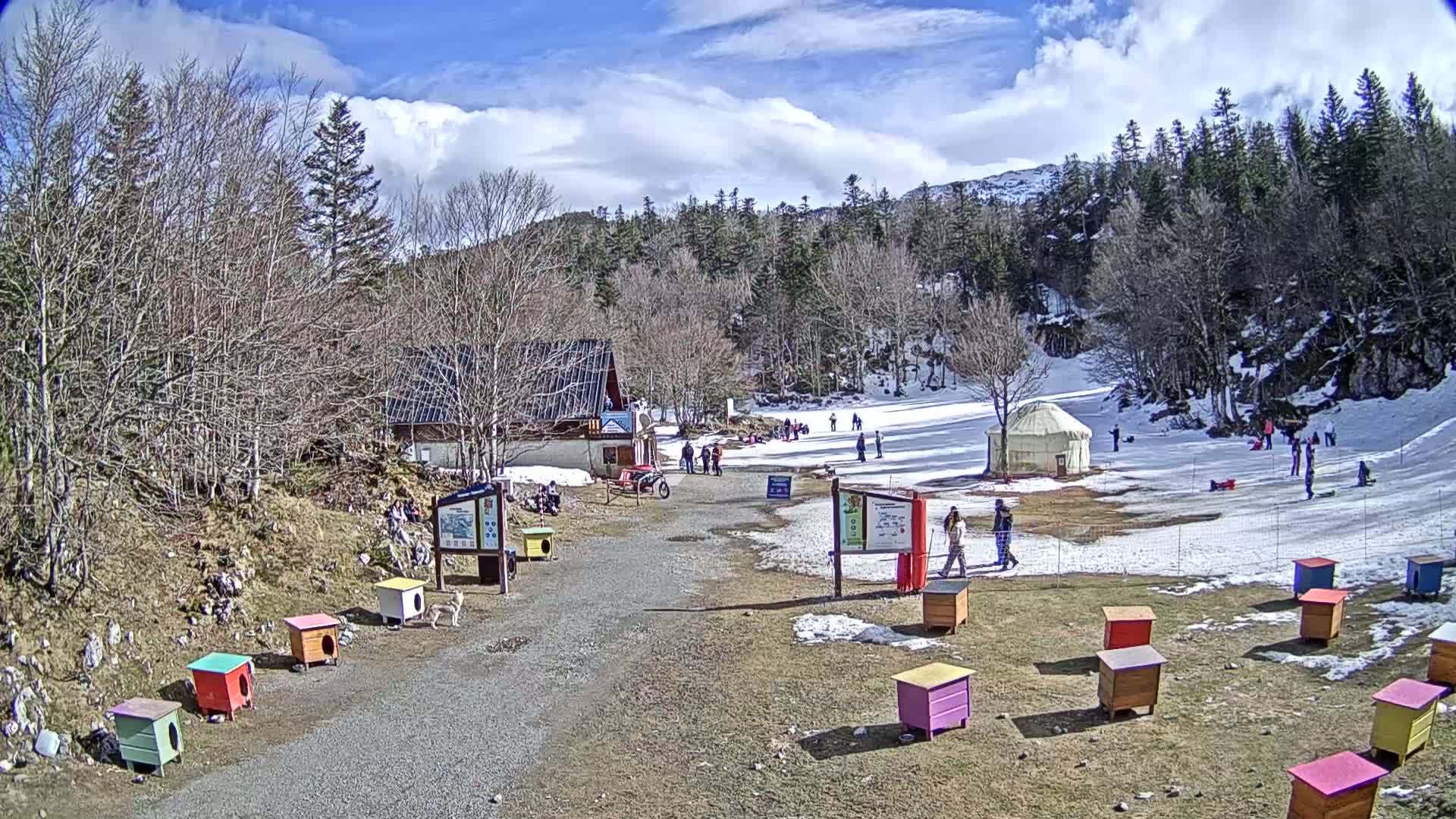 A sunny day at a snowy mountain resort shows a small ski slope with people enjoying activities, a yurt, a building, and several colorful dog houses.