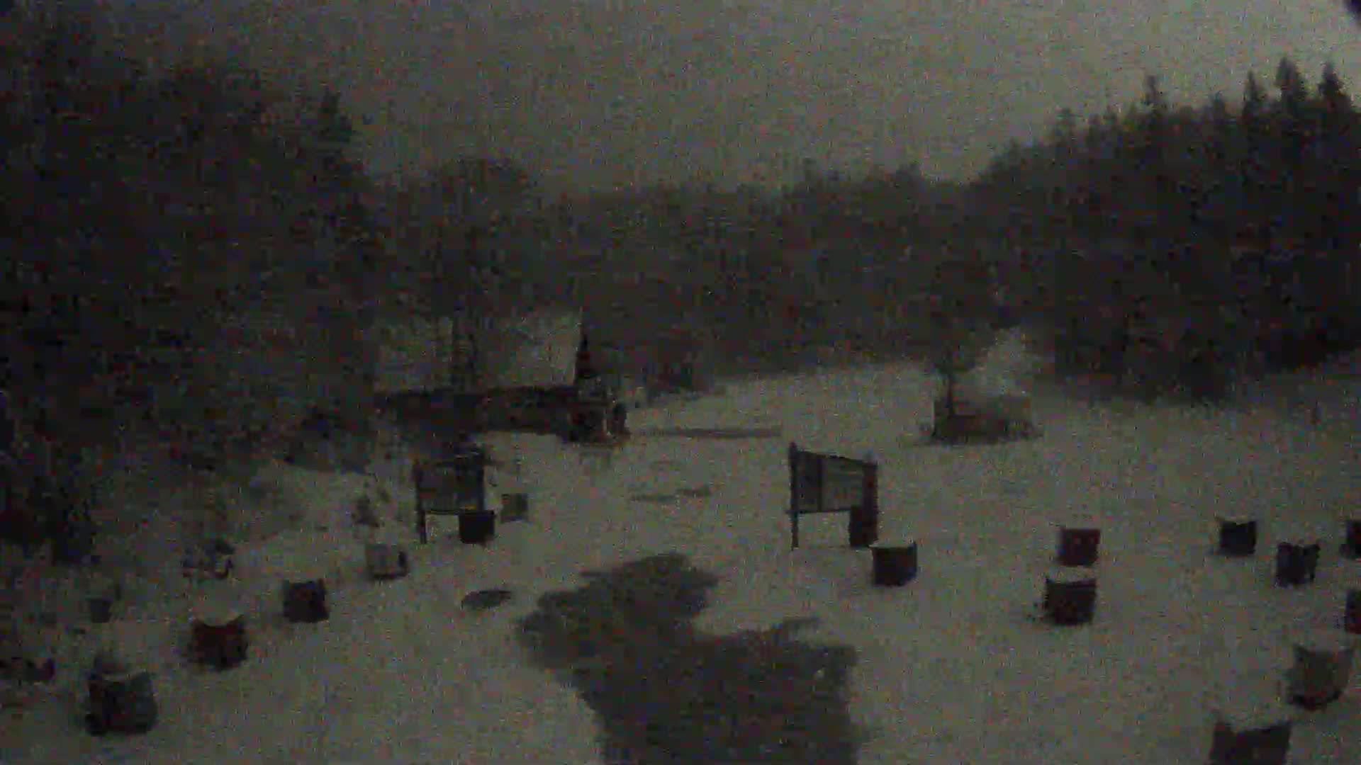 A snow-covered area with several dark rectangular objects scattered across it, surrounded by dark trees under a heavily overcast sky during a snowfall.