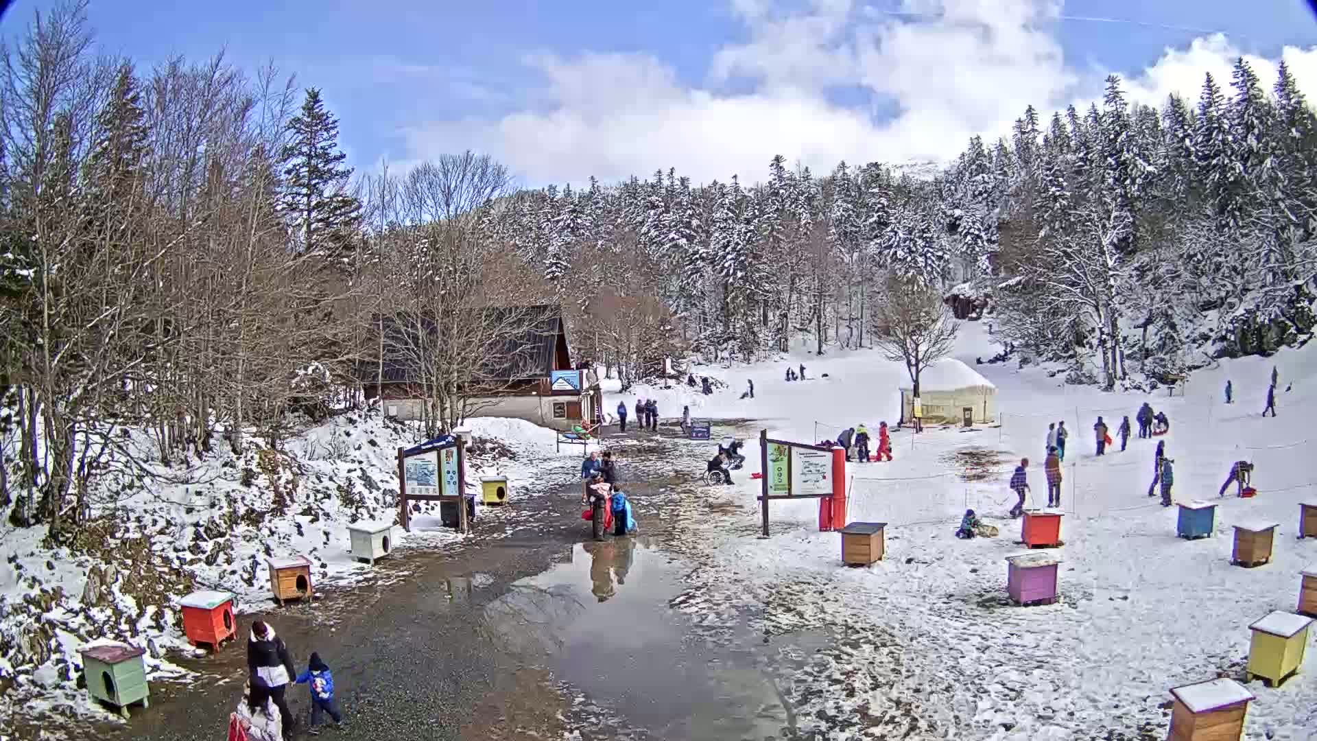 A snowy, sunny day at a small ski slope shows people sledding and walking near a building, surrounded by snow-covered trees.