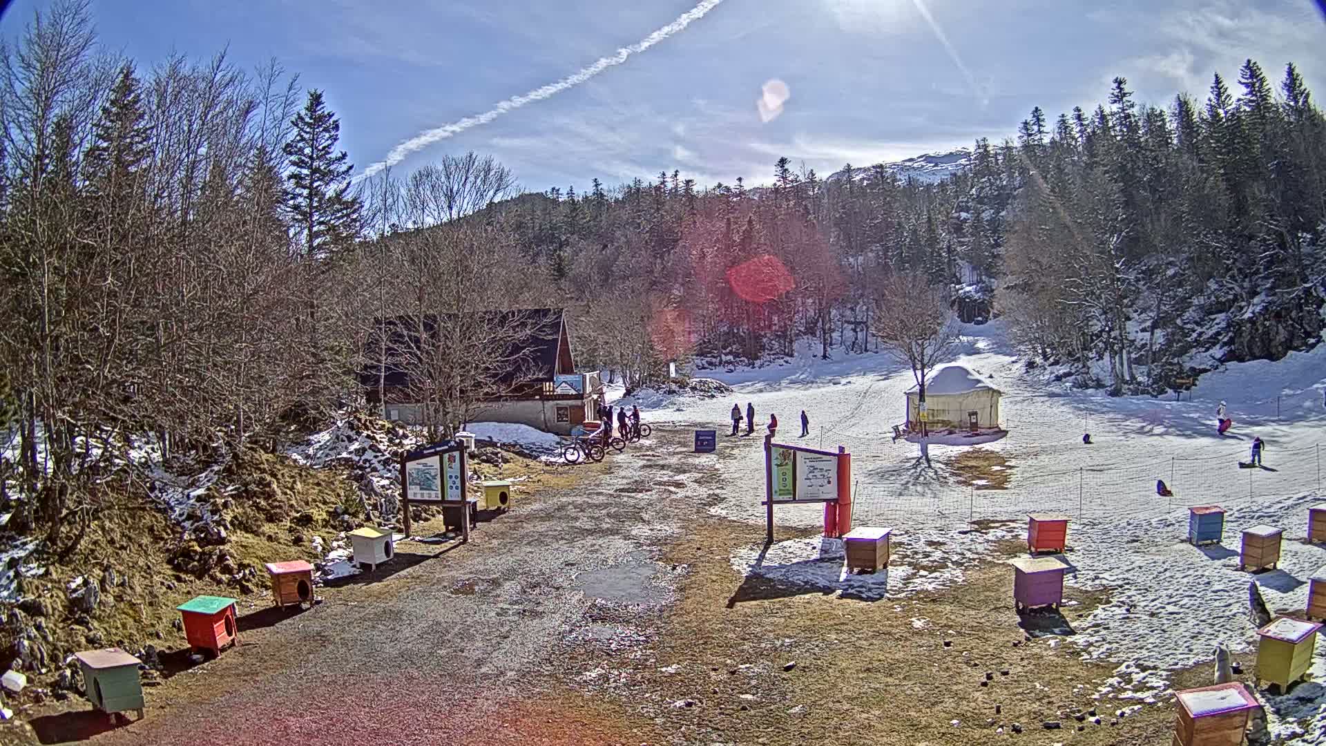 A sunny day at a snow-covered mountain resort shows a small building, a few people, several colorful structures, and numerous bare deciduous trees surrounding a snow-covered hill.