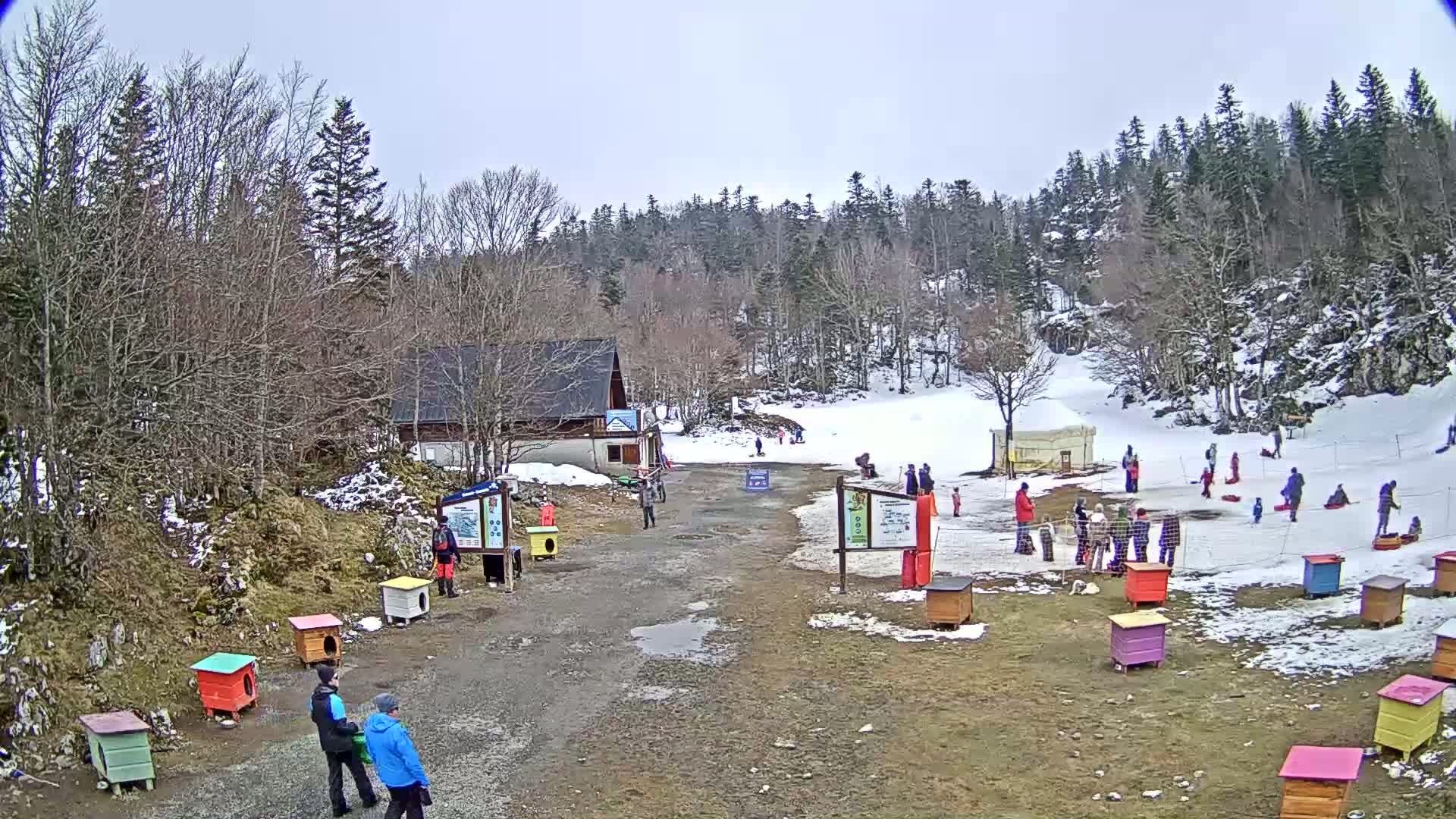 A snowy hillside with a small building, several people, and colorful wooden structures, under an overcast sky.