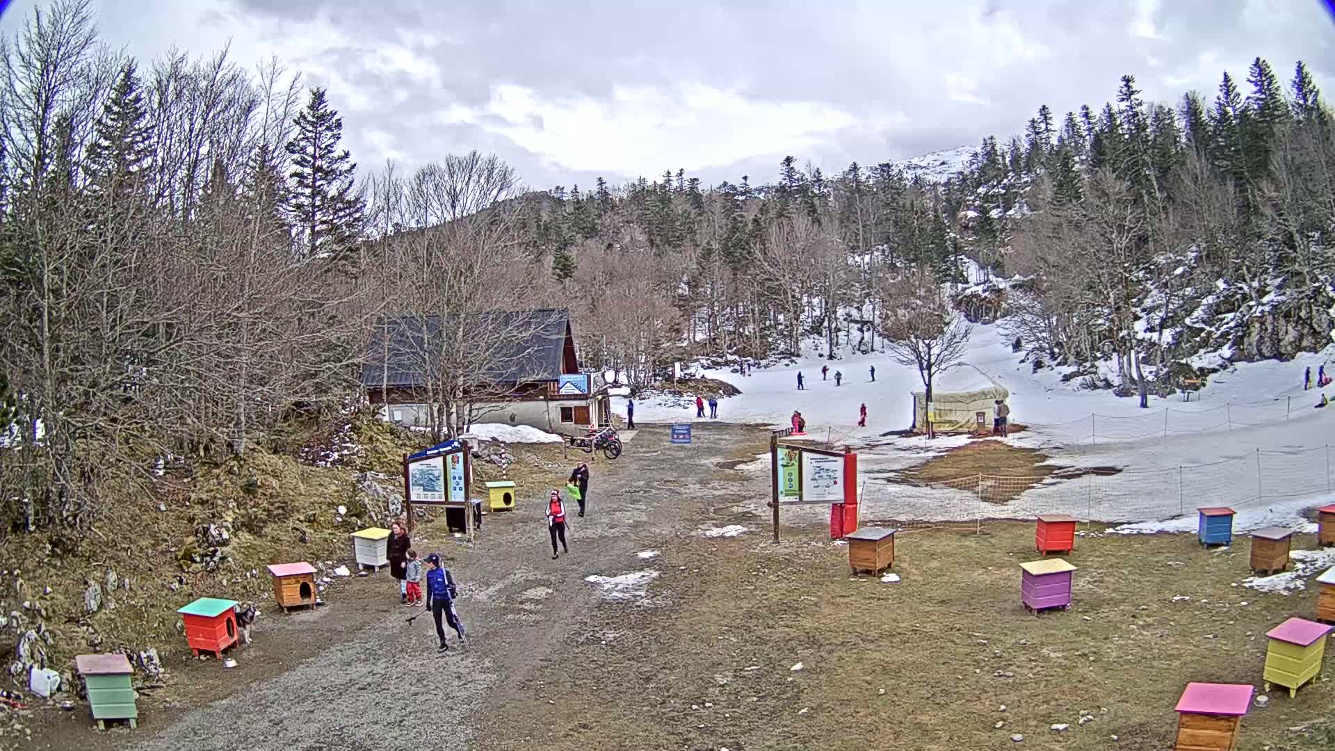 A partially snow-covered ski slope with several people, a small building, and colorful dog houses under a cloudy sky.