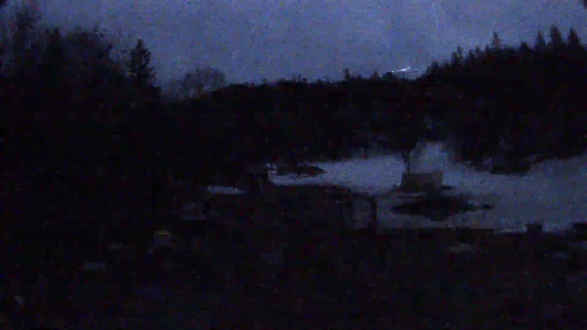 A dark, nighttime scene shows a snow-covered area with trees and buildings in the background under a dusky sky.