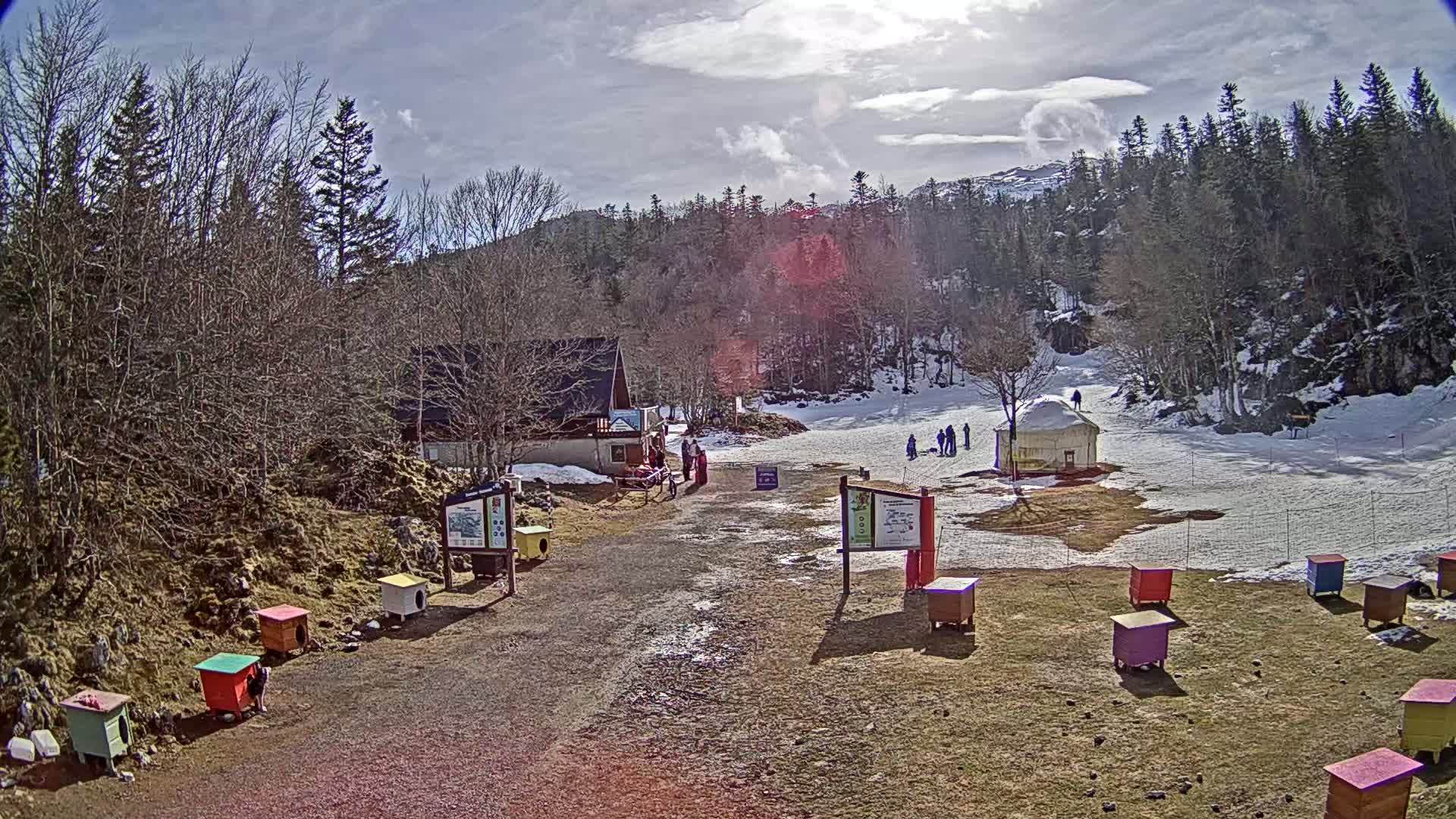 A sunny day at a snow-covered ski area shows a small building, several people, and numerous colorful structures near a dirt path.