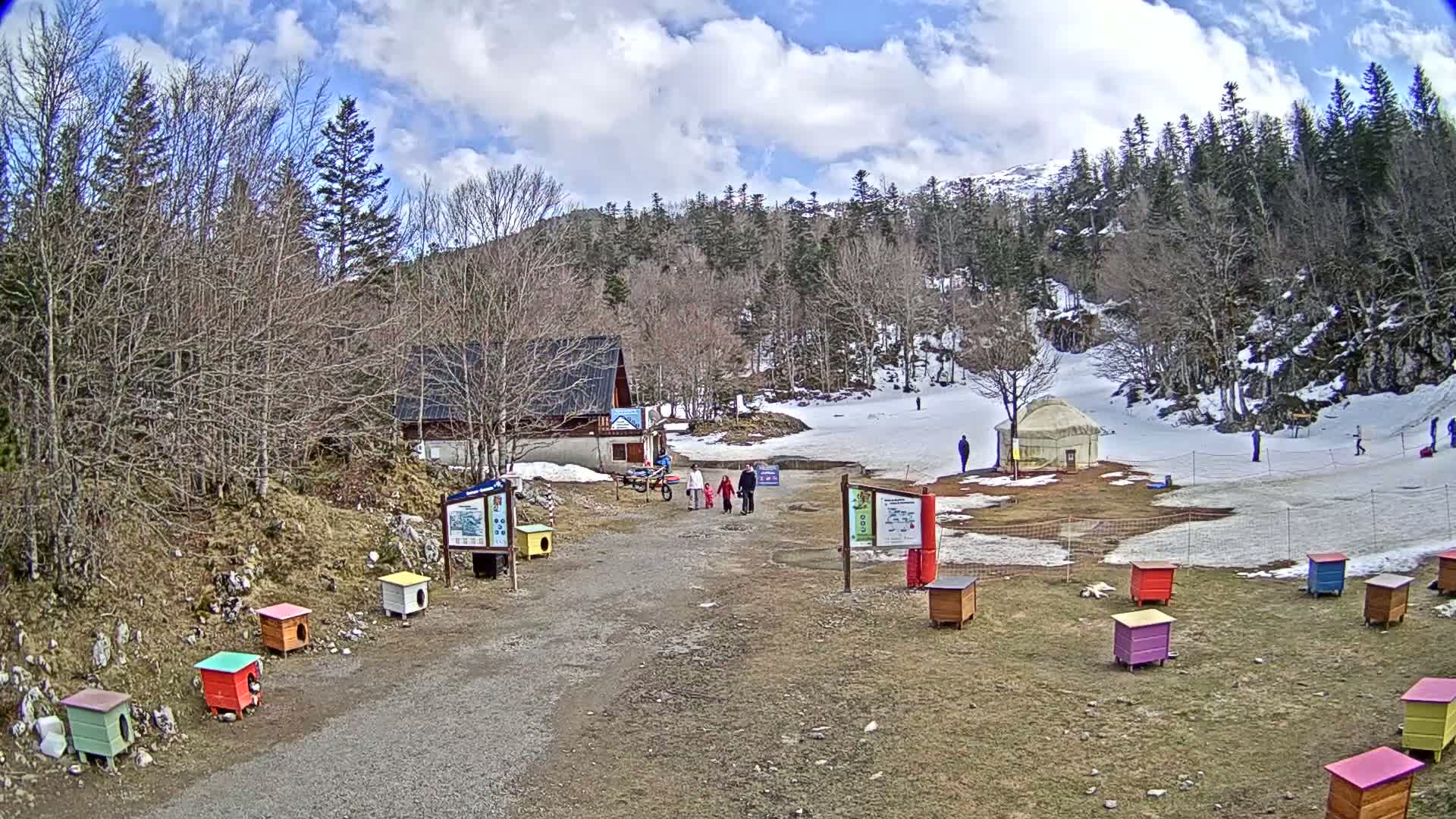 A partly sunny day at a snowy mountain resort shows a small building, a yurt, several colorful dog houses, and a few people walking on a gravel path.
