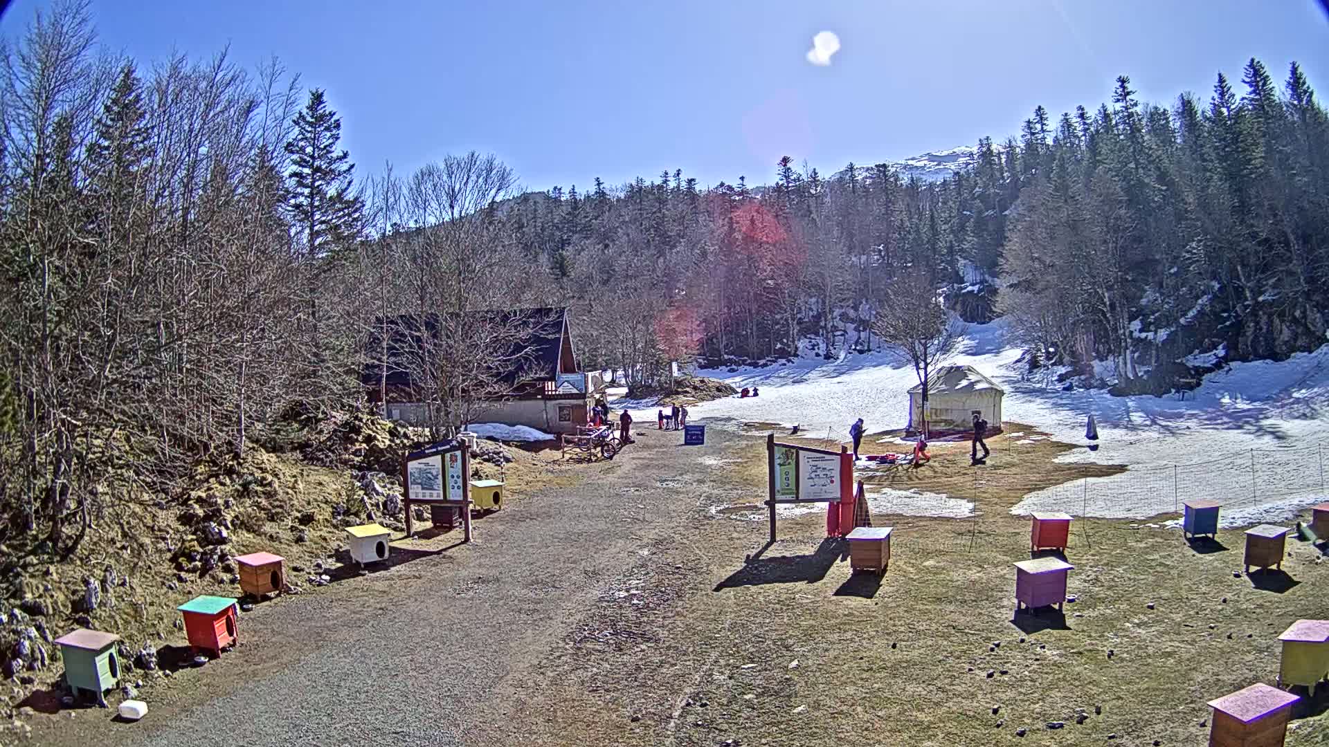 A sunny day at a small ski resort with a few people present, showing a building, a snow-covered slope, and numerous colorful small structures scattered on the ground.