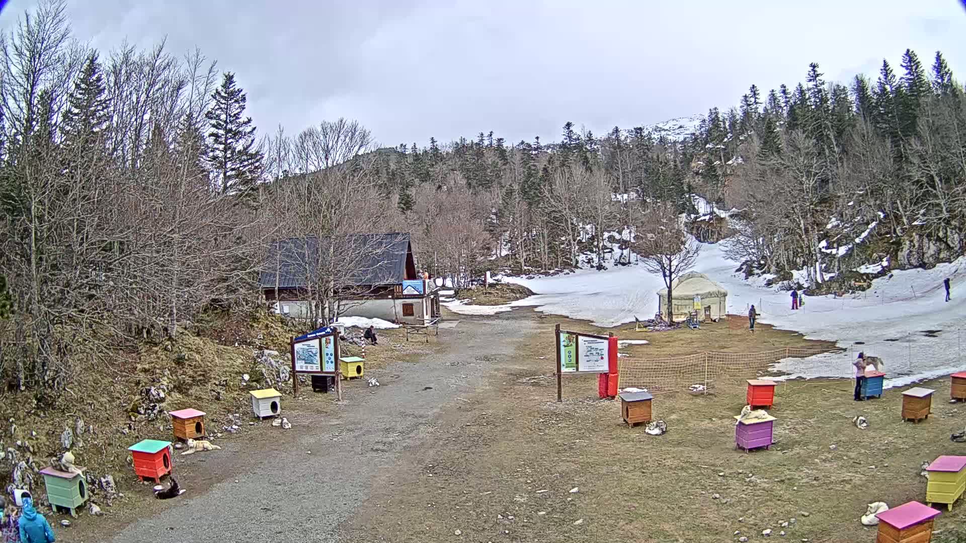 A snowy mountainside scene with a small building, a yurt, several colorful dog houses, and people, under an overcast sky.