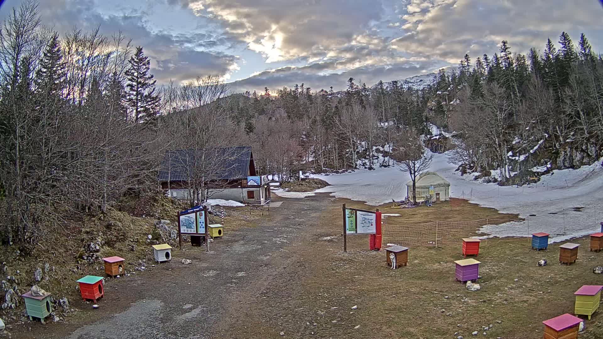 A mostly cloudy day reveals a mountainous landscape with a small building, a yurt, and numerous colorful dog houses scattered across a snow-dusted clearing.