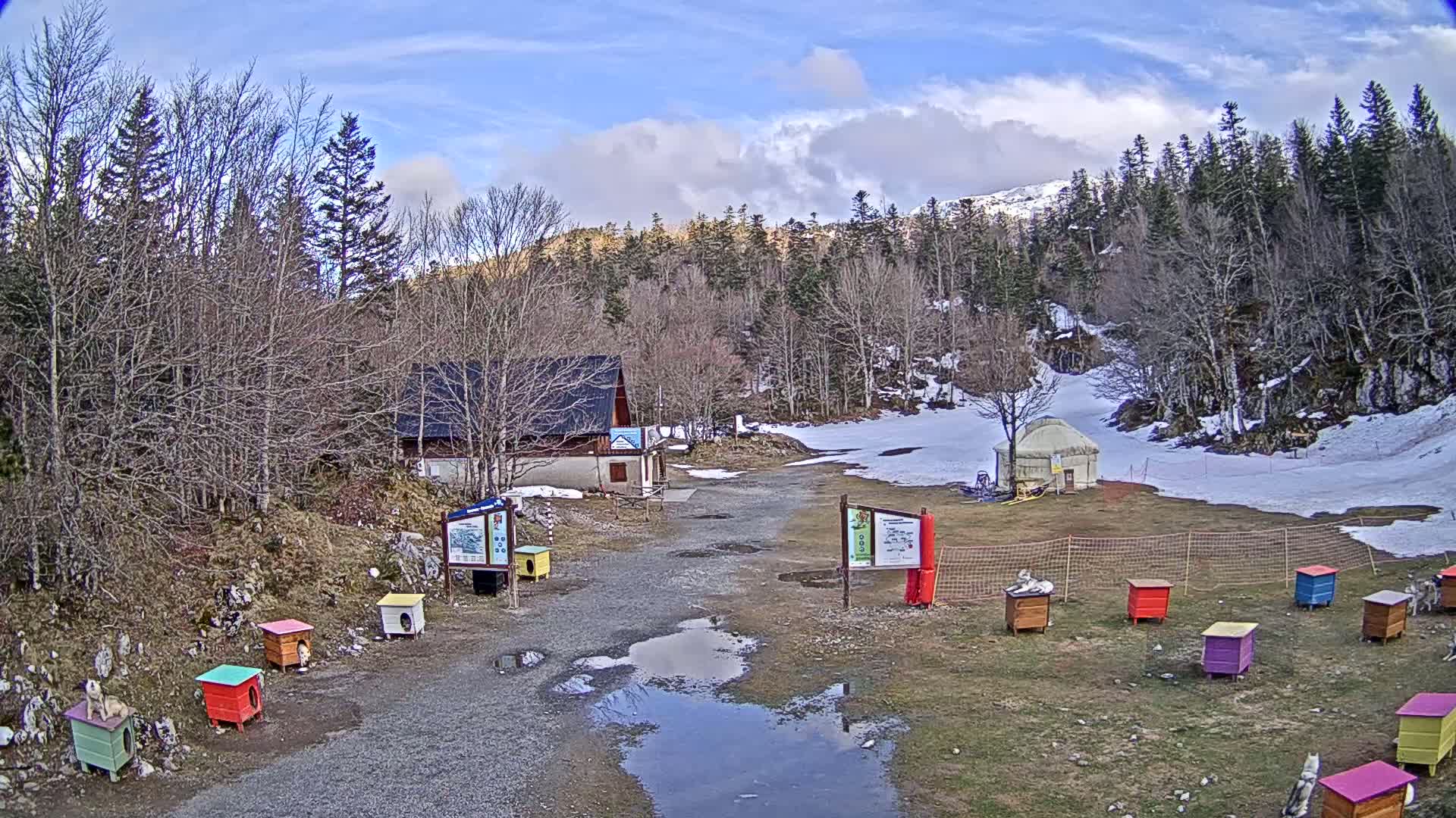 A small building sits near a yurt in a mountain area with patchy snow, under partly cloudy skies.