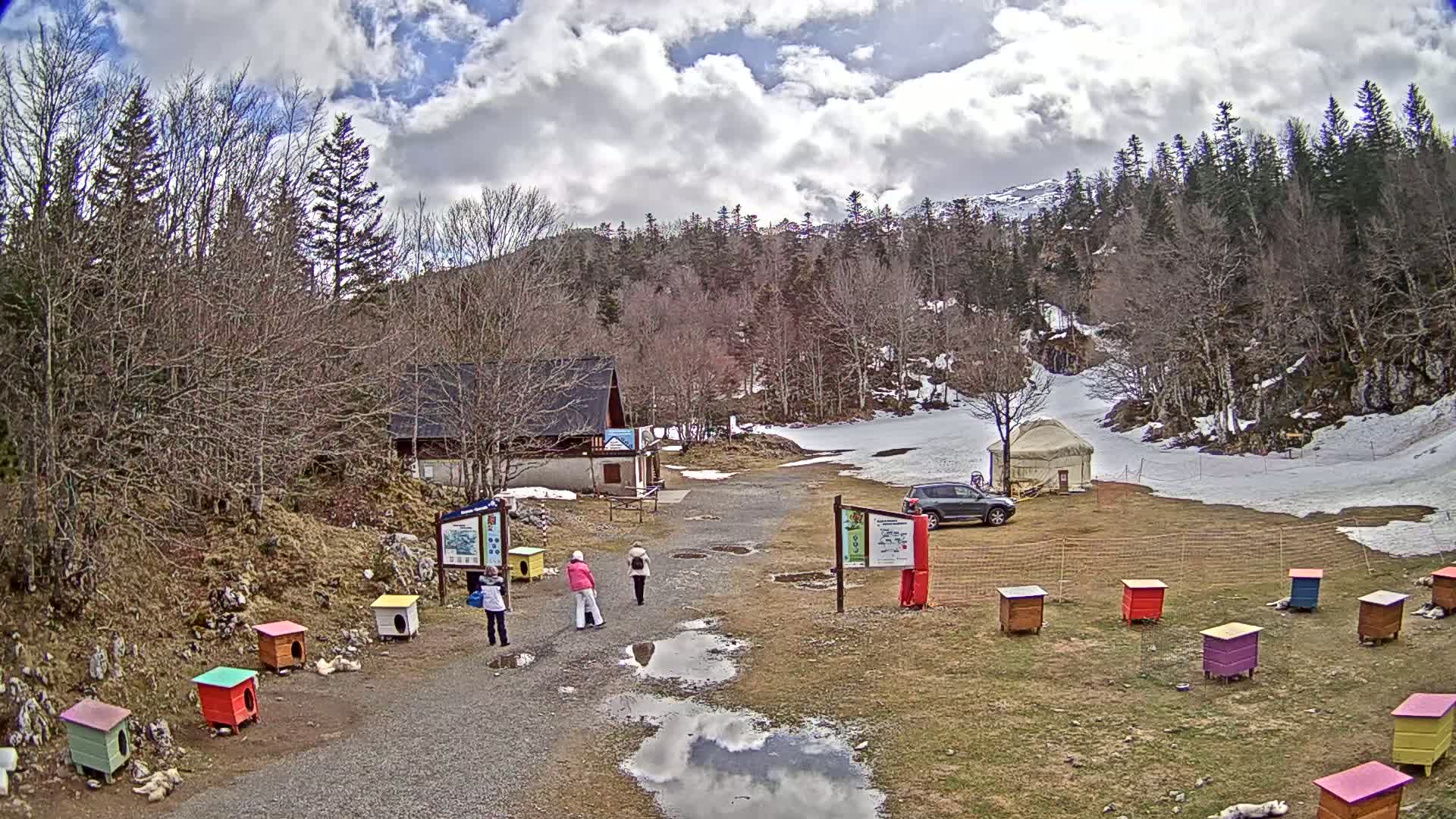 A mostly sunny day with patchy snow on the ground shows a parking area with several people, a yurt, a small building, and a line of colorful dog houses near a forest.