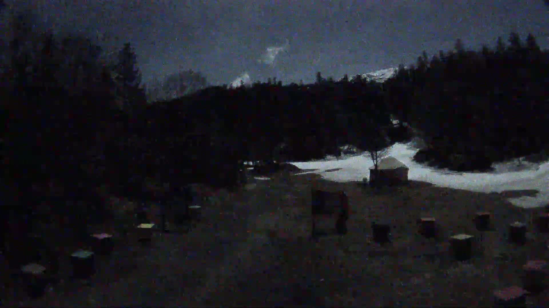 A dark, snowy nighttime scene shows a yurt and numerous small structures scattered in a clearing amongst dark trees under a cloudy sky.