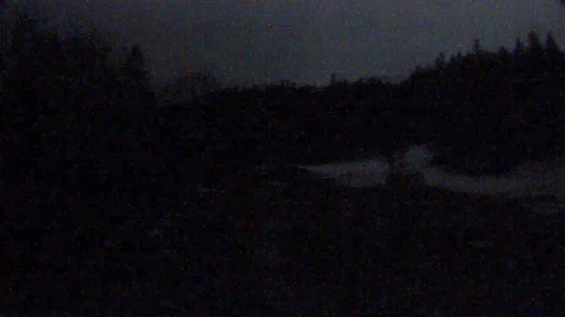 A dark, nighttime scene shows a body of water partially visible behind a dark, wooded area under a dusky sky.