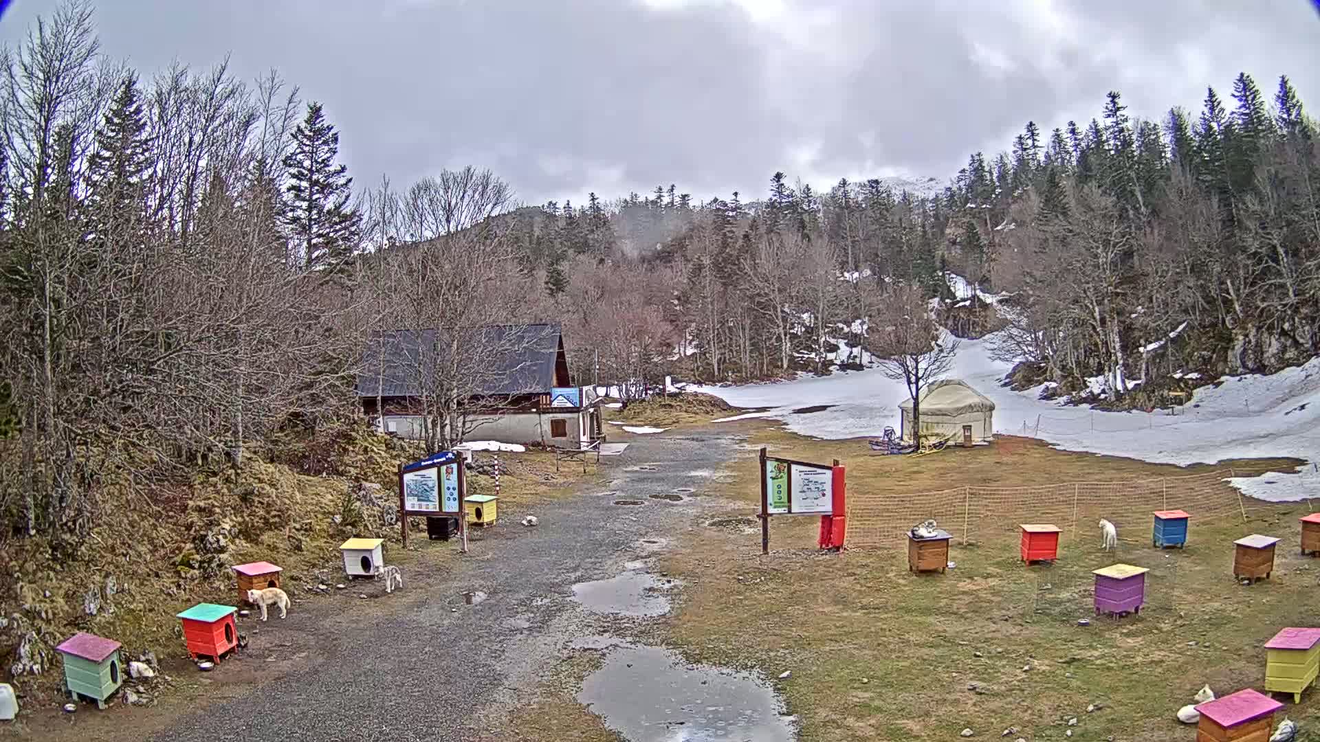A gravel path leads to a small building and a yurt in a mountainous area with patchy snow on the ground under an overcast sky.
