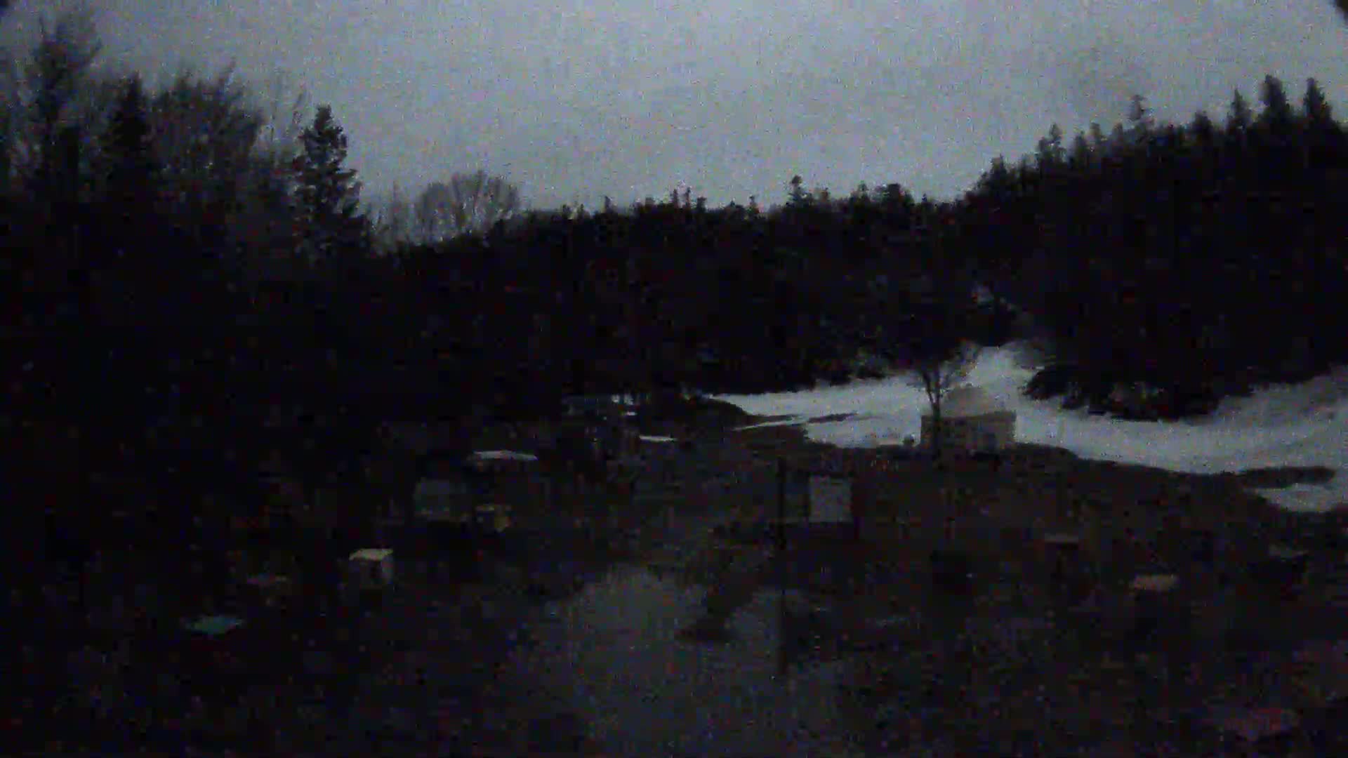 A dark, nighttime scene shows a snow-covered area with a few structures and trees under a gray, overcast sky.