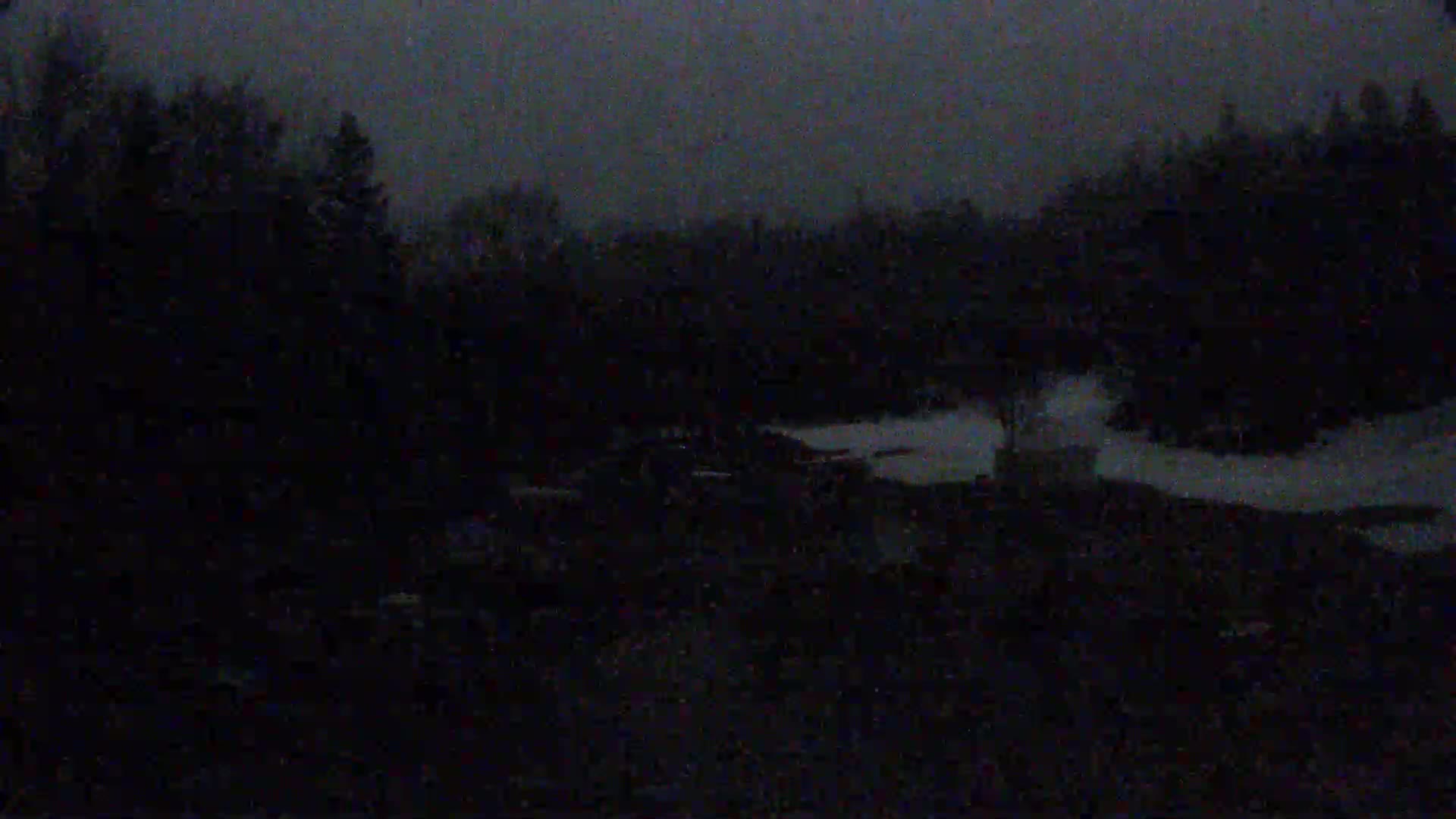 A dark, nighttime scene shows a body of water with a small structure near its edge, surrounded by trees under a dusky sky.