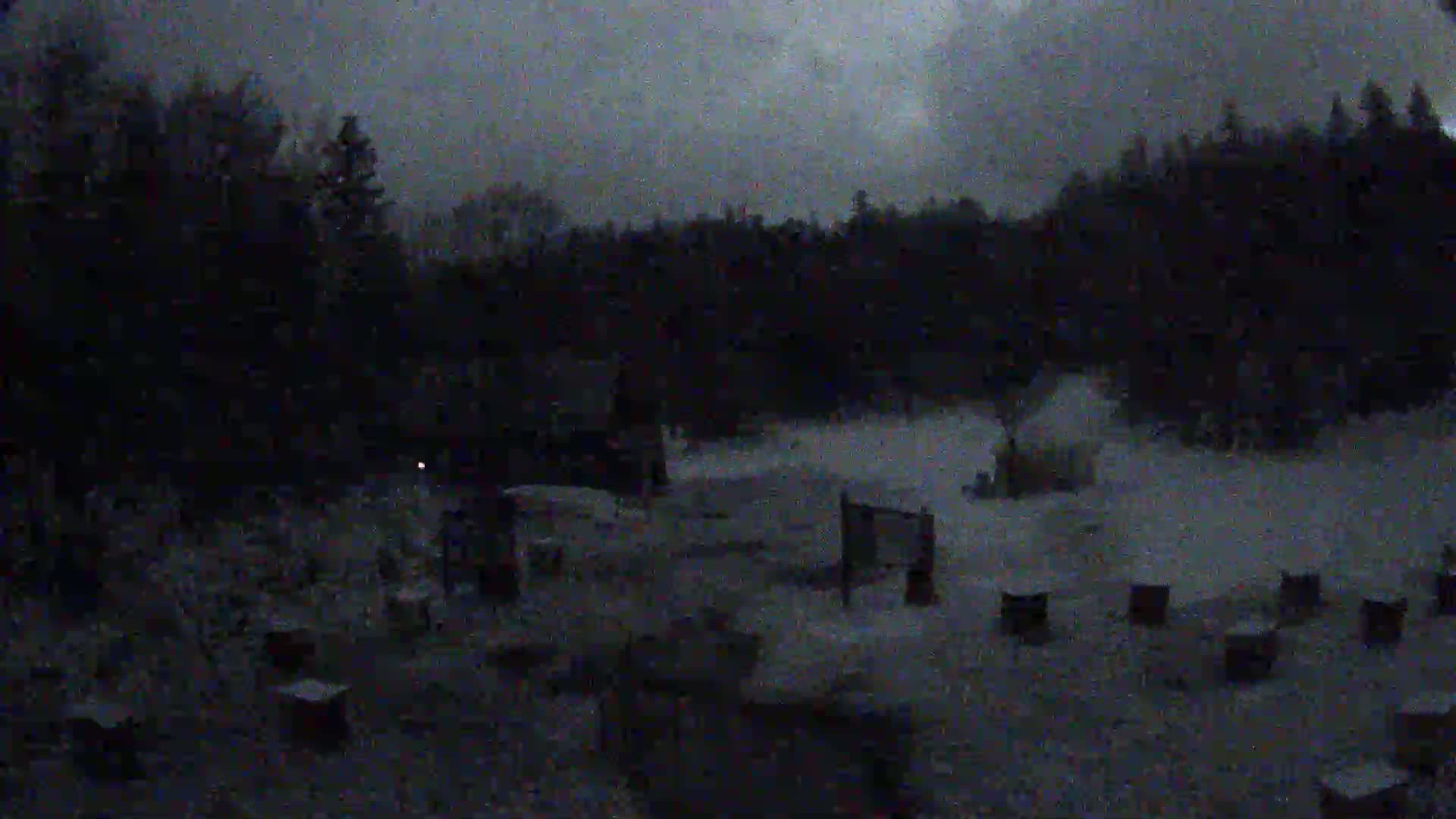 A snow-covered area with several small structures and a line of dark trees in the background under a dark, overcast sky.