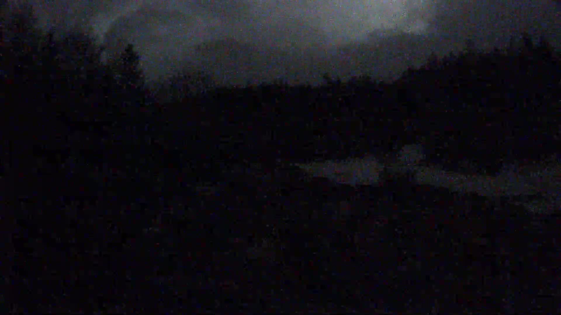 A dark, nighttime scene shows a body of water near a silhouetted line of trees under a stormy sky.