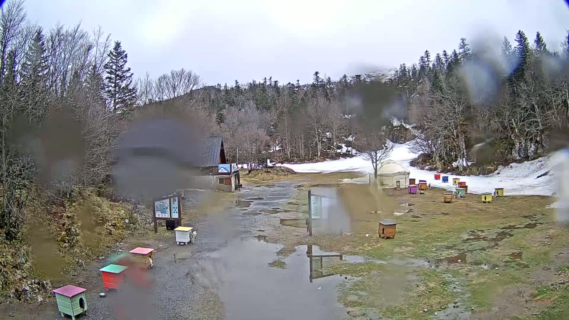 A rain-soaked, muddy area with several colorful small structures, a building, and a yurt sits in a clearing surrounded by a snowy, wooded hillside.