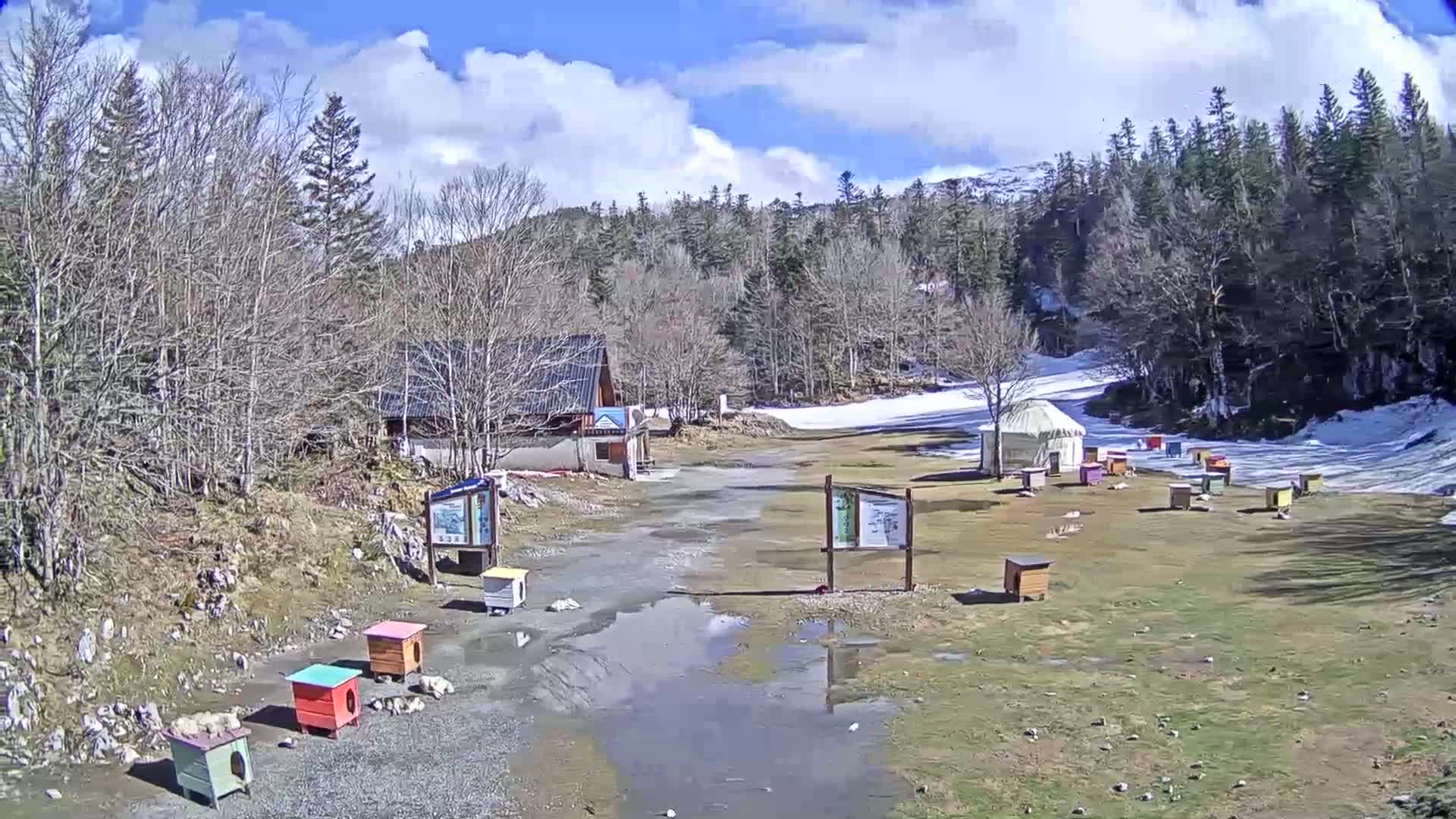 A partially snow-covered landscape with several small, colorful structures, a yurt, a building, and puddles under a partly cloudy sky.
