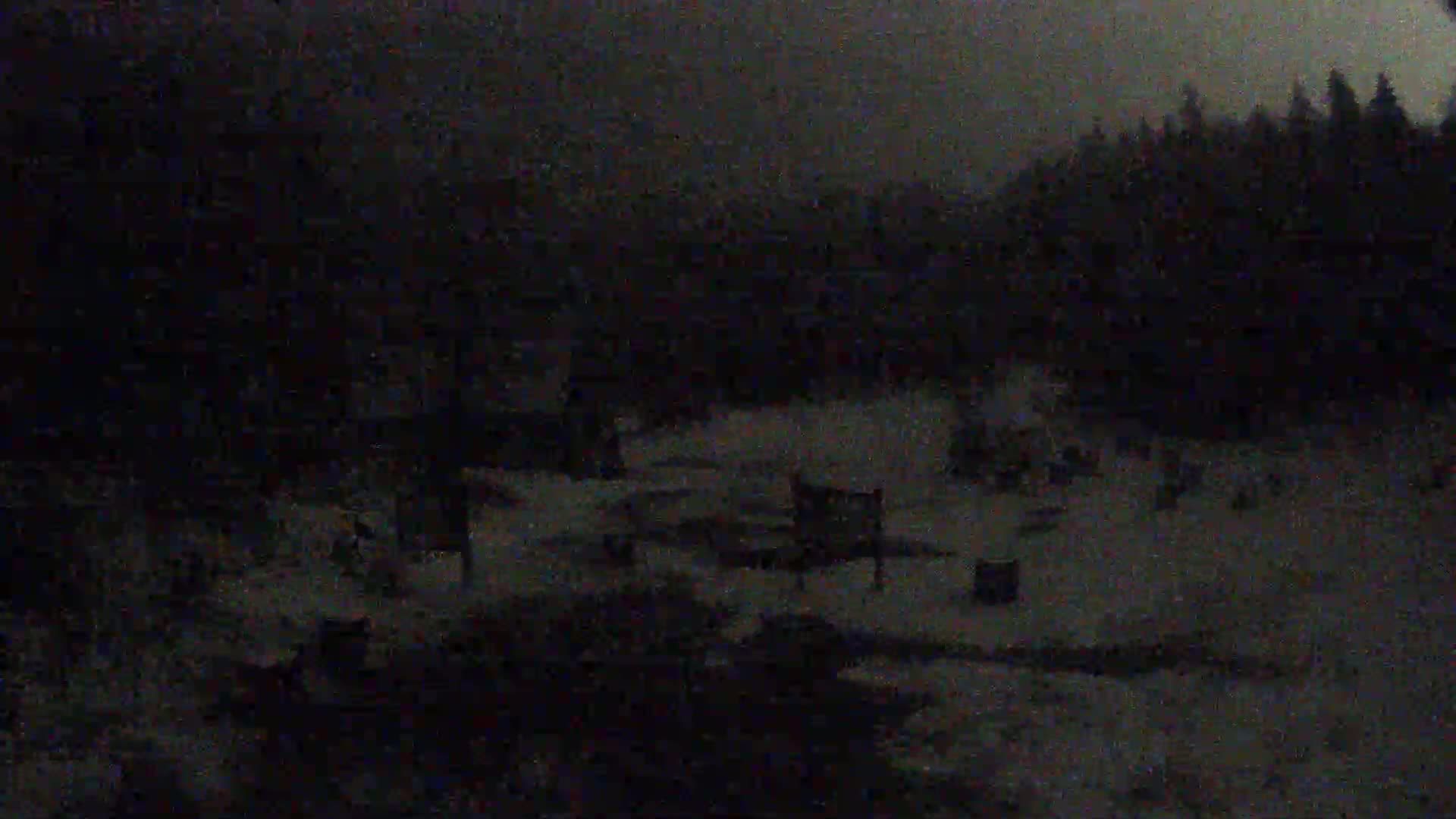 A dark, snowy nighttime scene shows a sparsely populated area with a few structures and trees in the background.