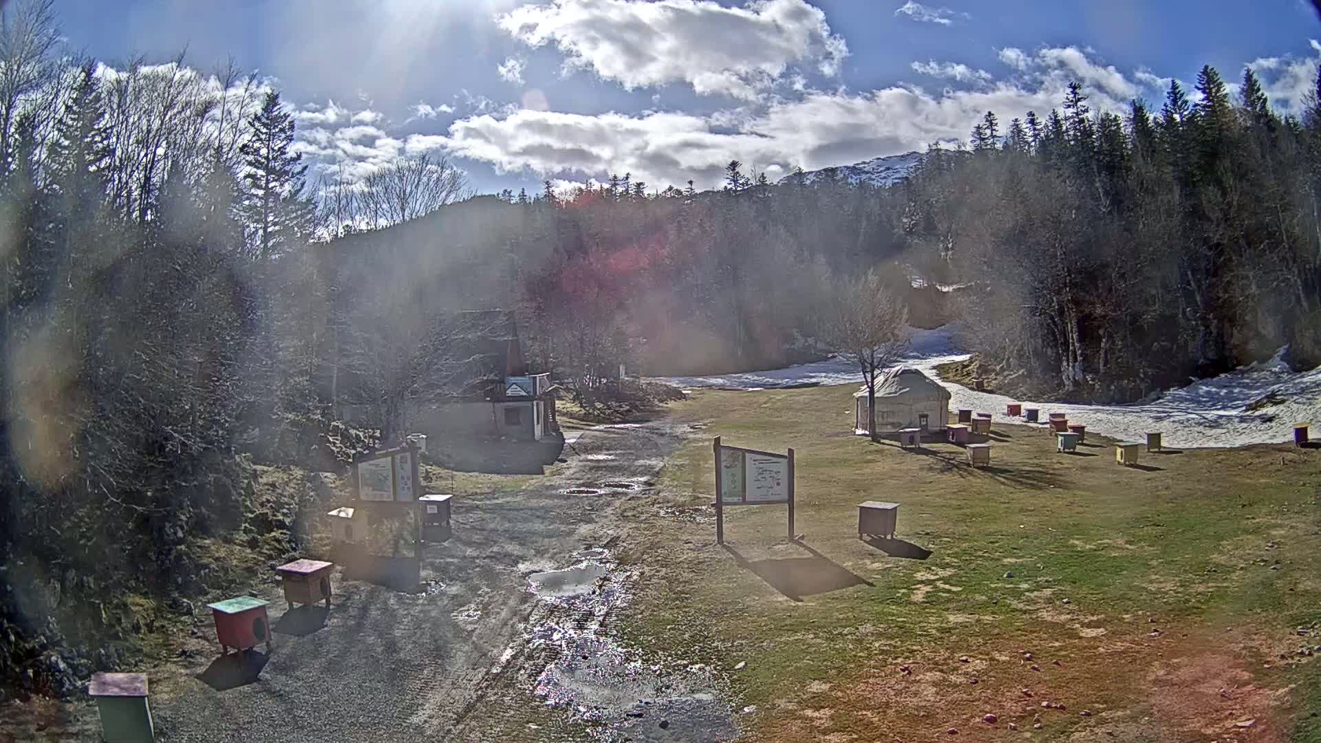 A sunny day with partly cloudy skies shows a dirt road leading to a grassy area with several small structures and a yurt, surrounded by a forest with patches of snow.